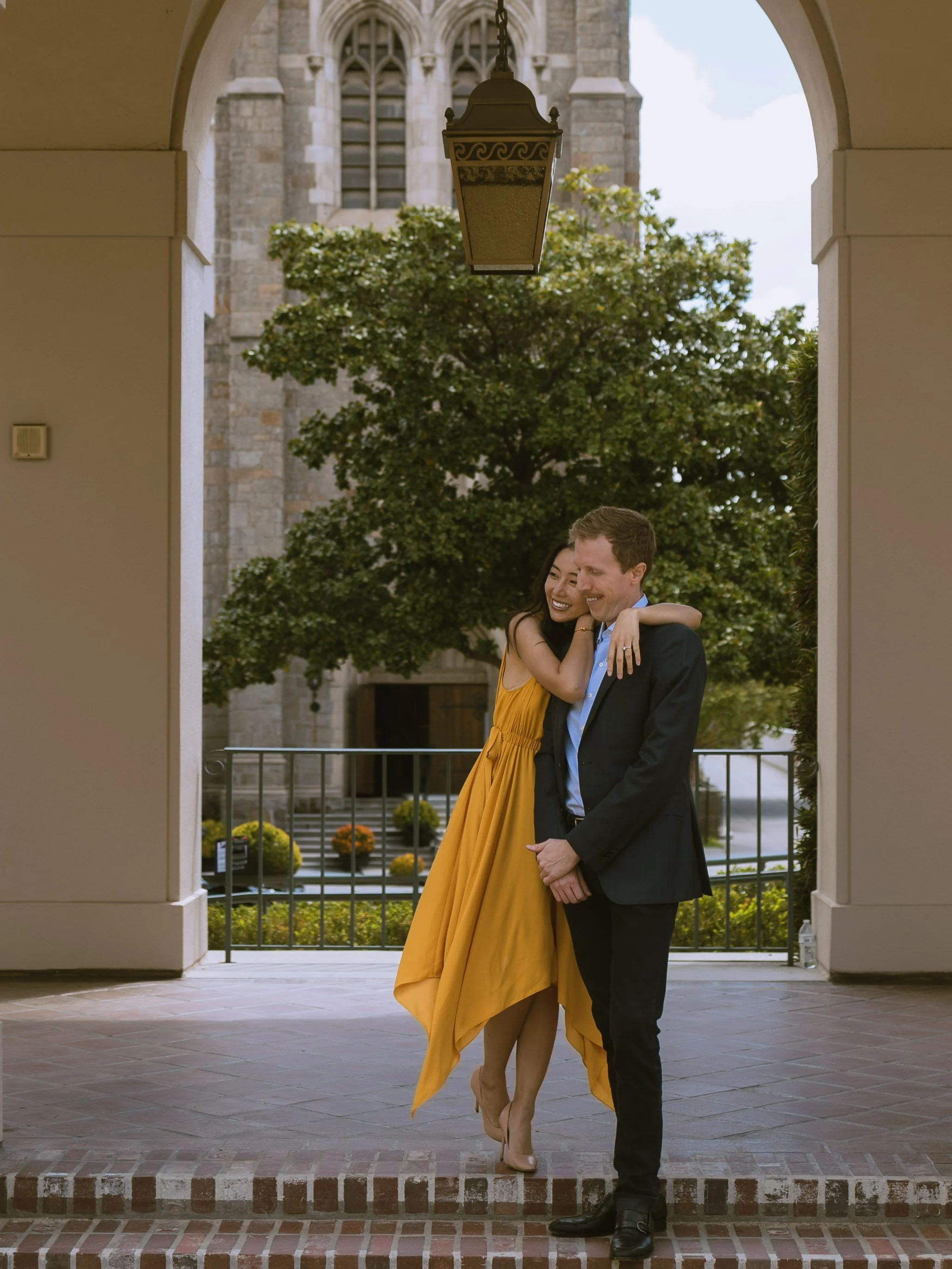 A couple is standing under an archway in front of a church, smiling and hugging. The woman is wearing a long yellow dress and heels, and the man is dressed in a suit and tie. There is a church tower and trees in the background, with potted flowers an