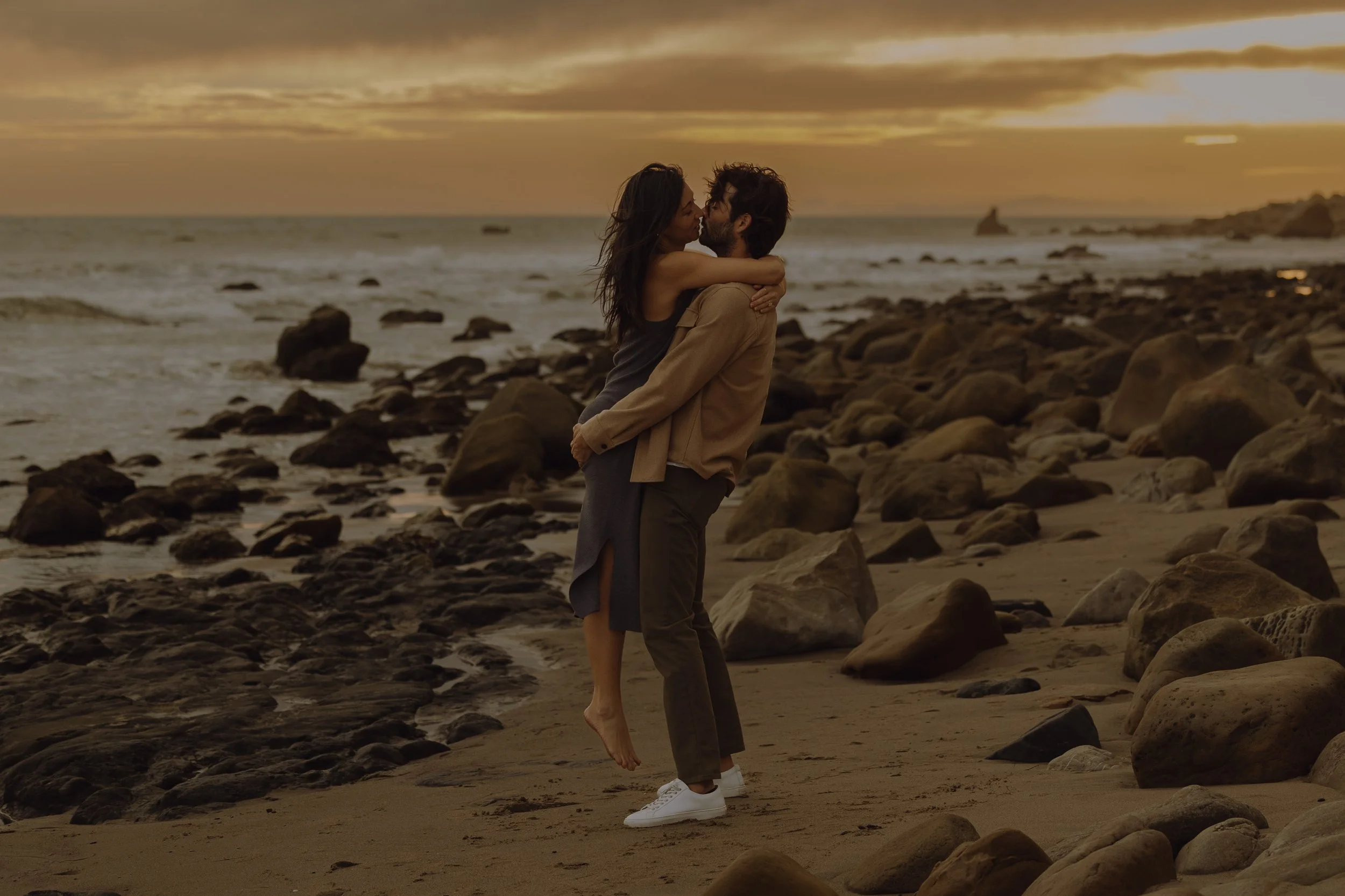 Couple embracing and kissing on a rocky beach at sunset, with the woman being lifted slightly by the man, both barefoot, with clouds and ocean in the background.