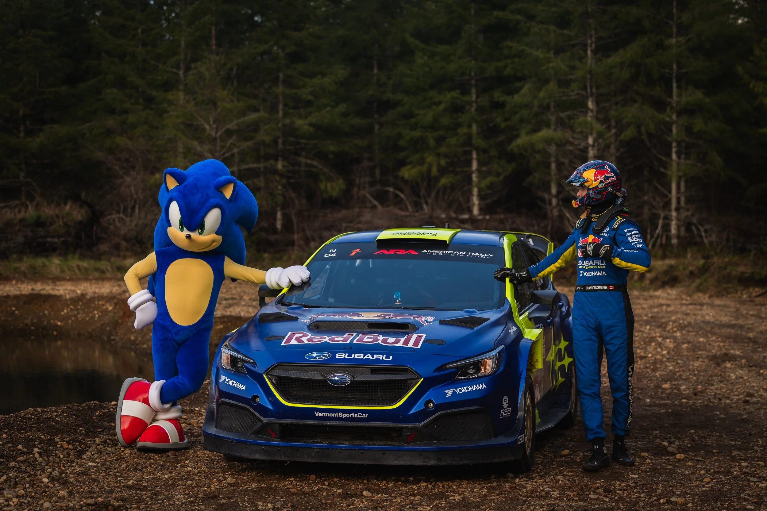 Sonic the Hedgehog mascot standing next to a rally car with a driver in racing gear, outdoors with a forest background.