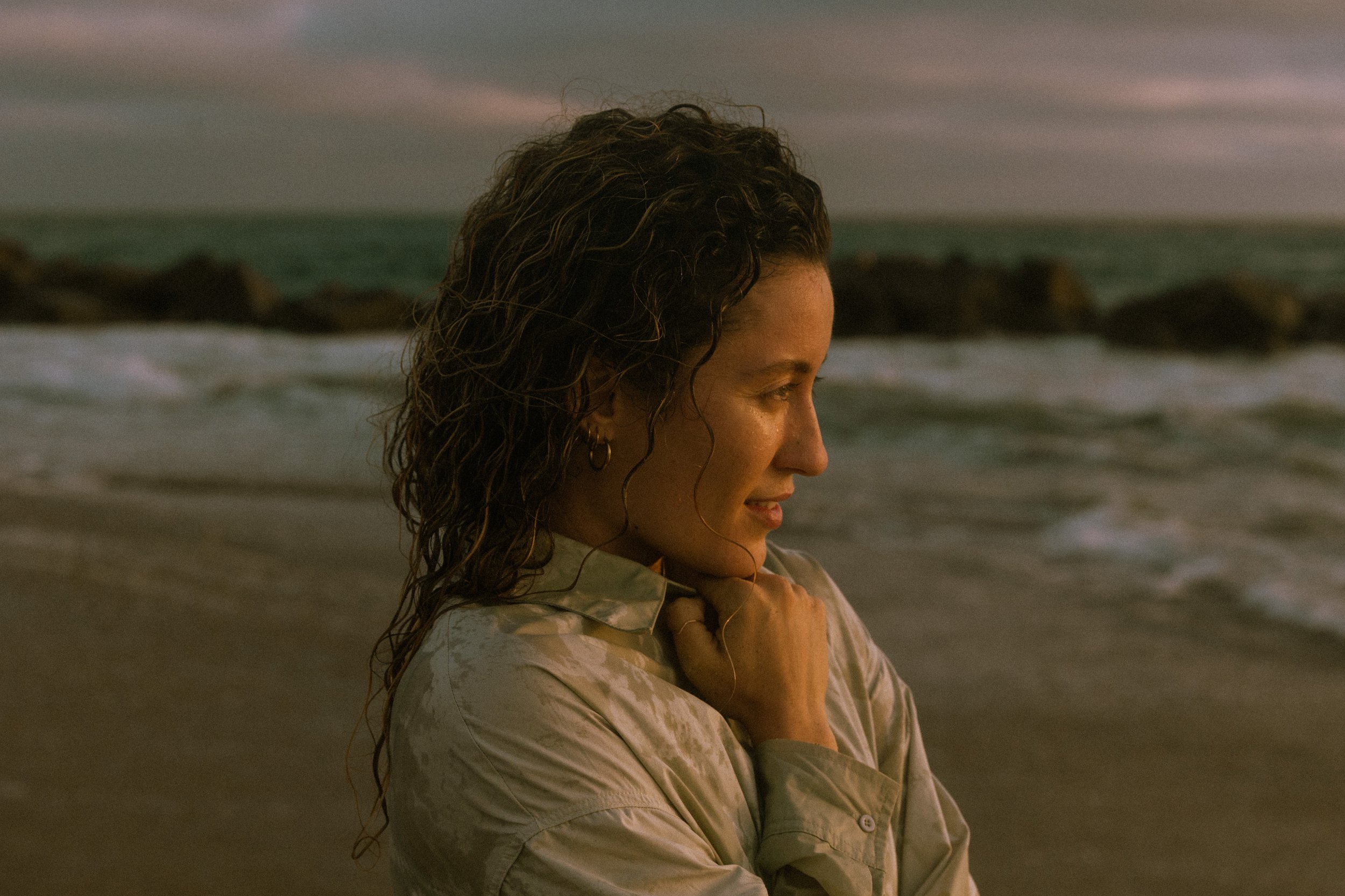 A woman with curly hair and earrings standing on the beach at sunset with her arms crossed and a serene expression.