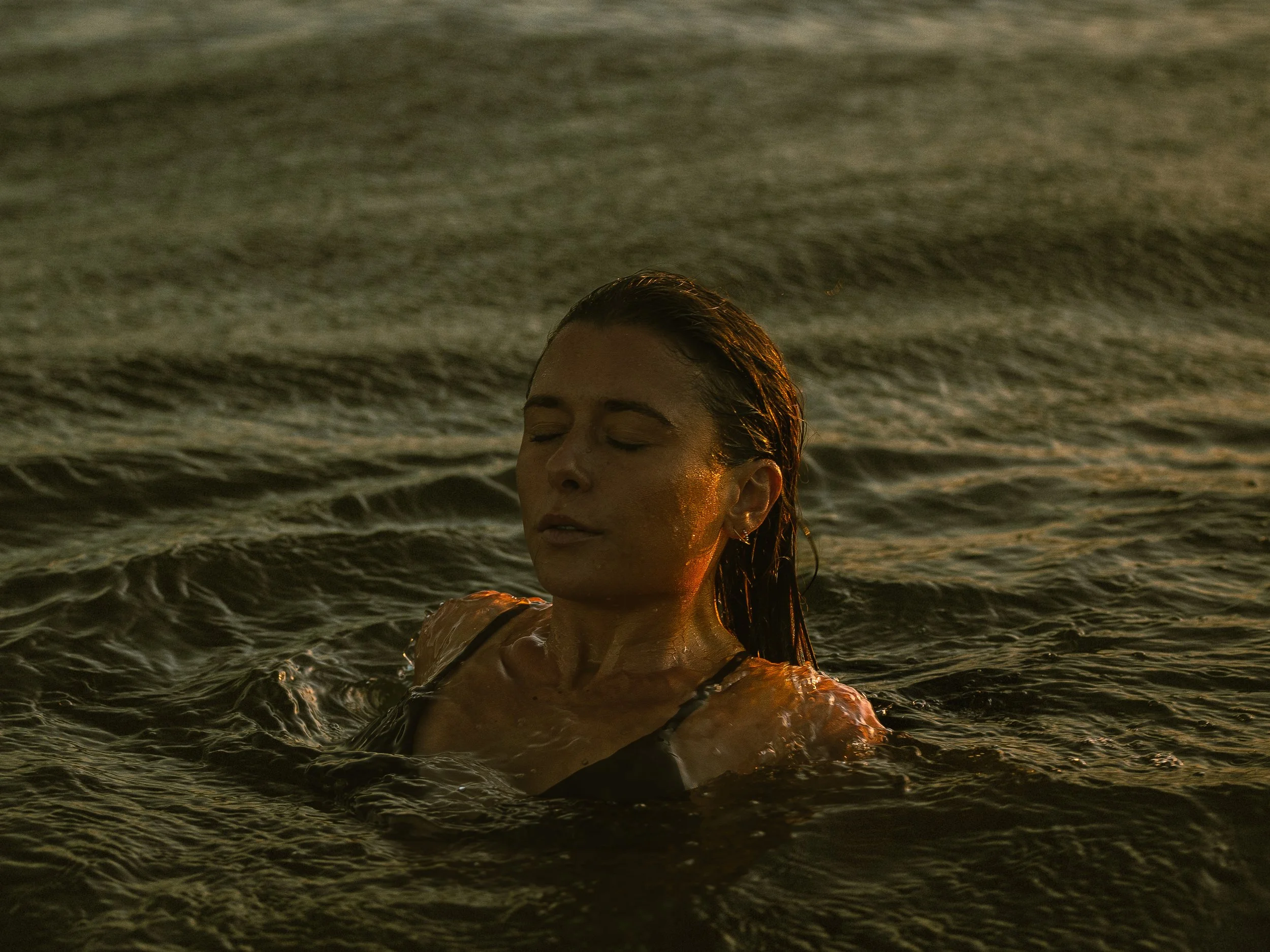 A woman with wet hair and closed eyes is in the water at sunset, with waves around her.