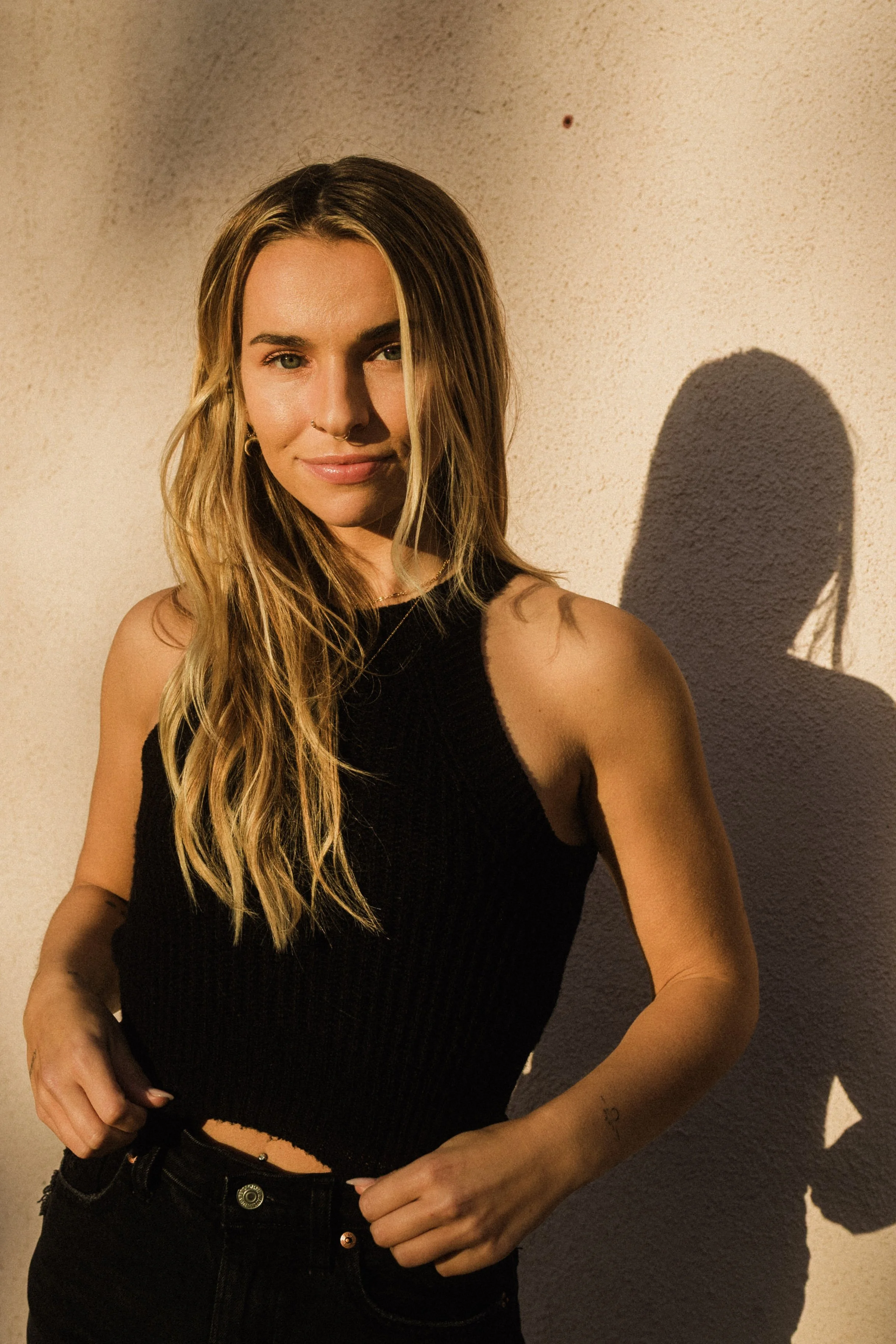A young woman with long blonde hair, wearing a black sleeveless top and black jeans, standing against a beige wall with the shadow of her head cast behind her.