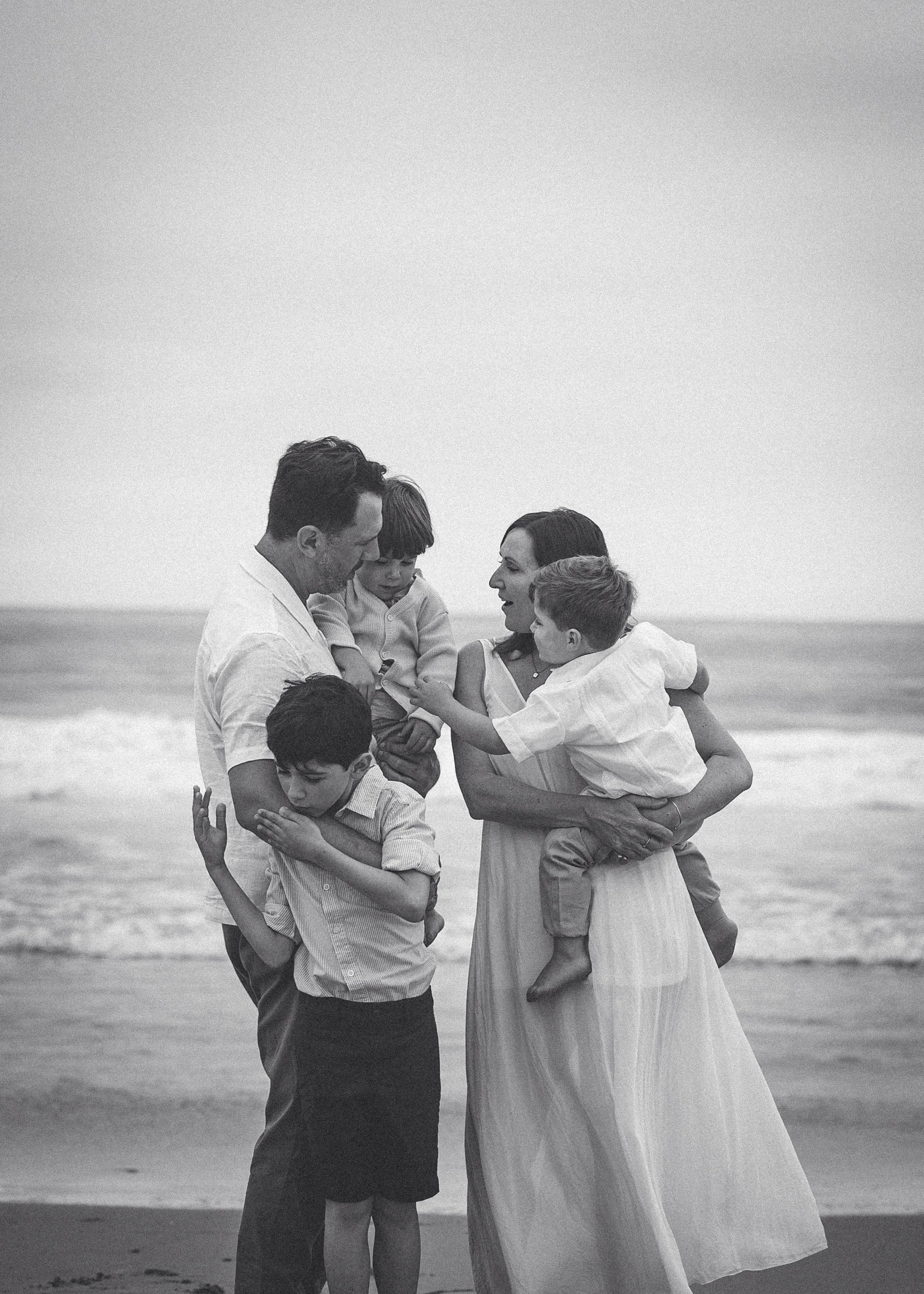 A family of five at the beach, sharing a moment of laughter and affection. The family includes two adults and three children. The background shows the ocean waves.