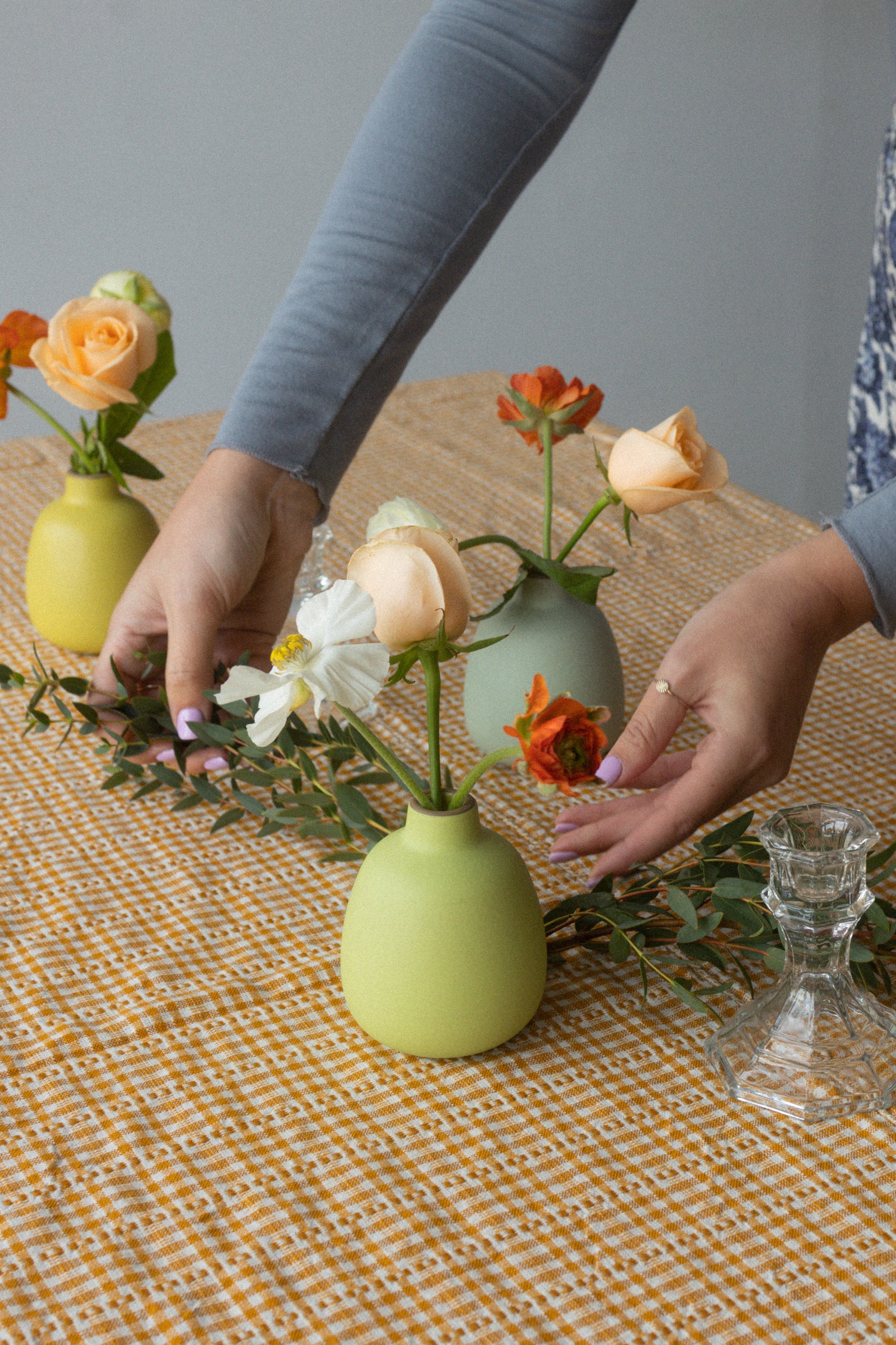 Hands arranging flowers in small green vases on table with orange and white checkered tablecloth, with glass candle holder nearby.
