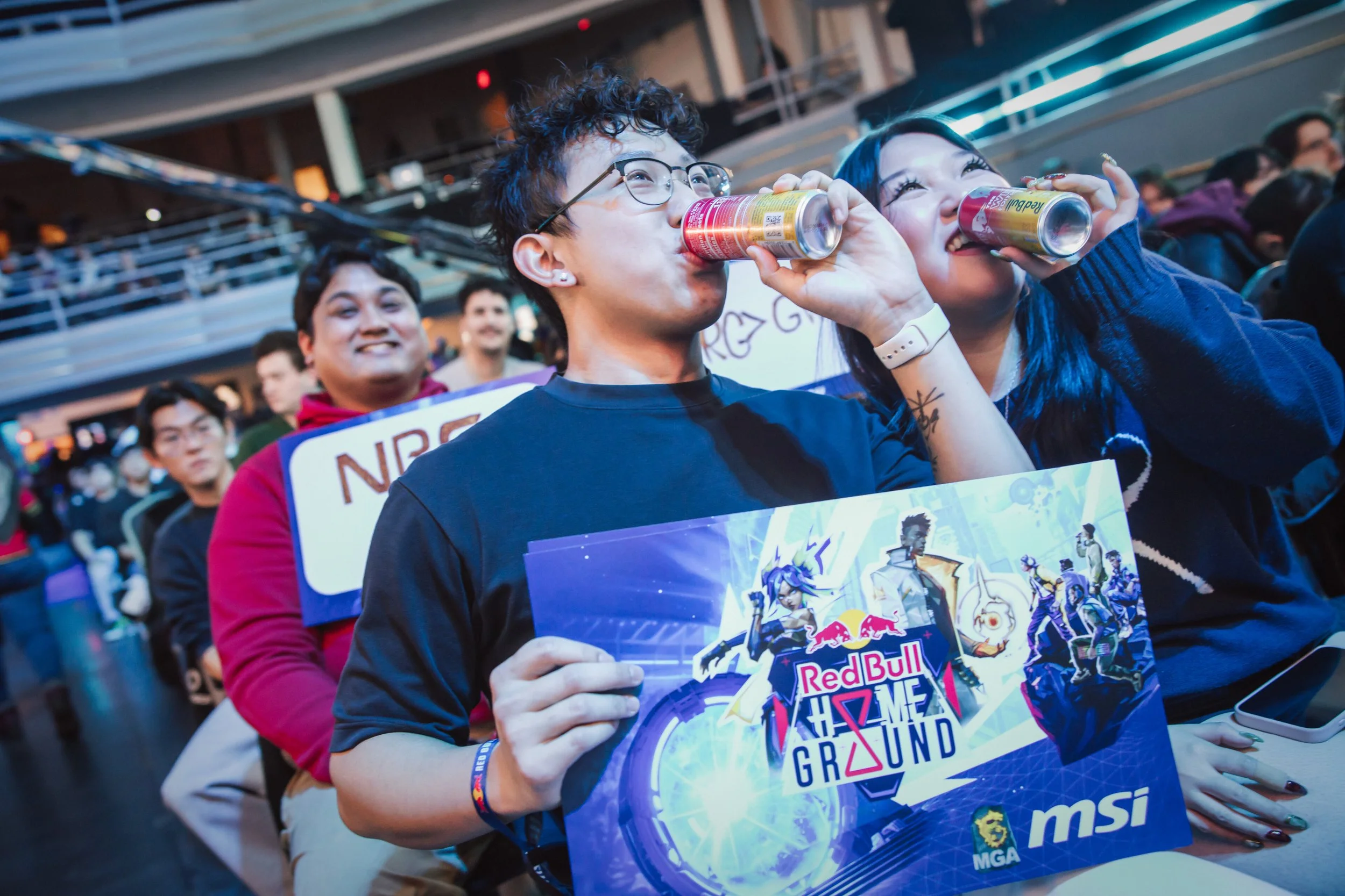 Two young people, a man with curly hair and glasses and a woman with dark hair, are drinking from Red Bull cans at an event. They are smiling and holding a colorful promotional sign for Red Bull gaming. There are other people in the background, and the setting appears to be an indoor arena or large venue.