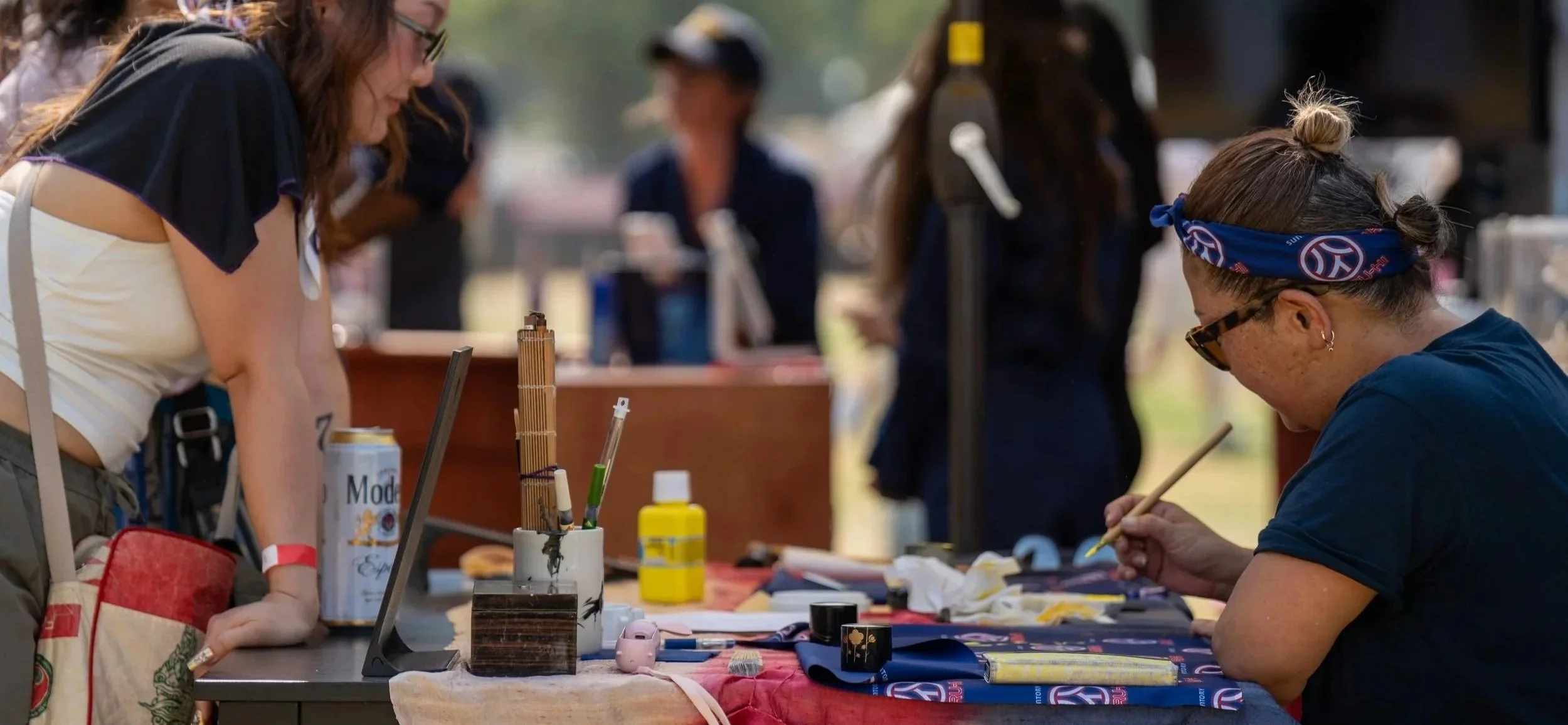 Two women at an outdoor table with art supplies, painting or crafting, with several people blurred in the background.