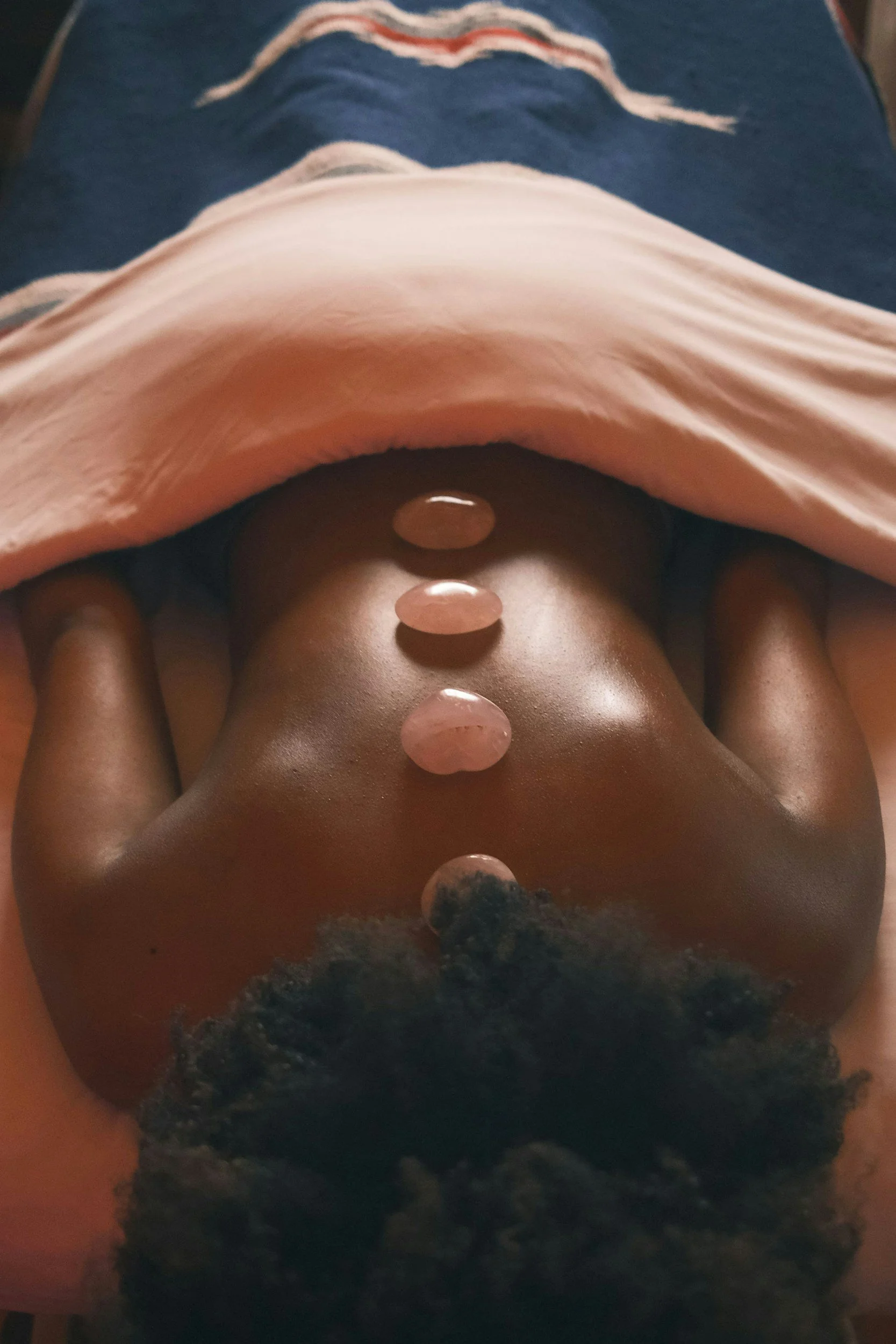 A person lying on their back on a blue surface, with a pink cloth draped over their lower body and small round stones placed along their abdomen.