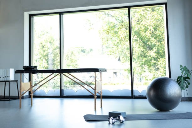 A bright, minimalist exercise room with large glass sliding doors showing greenery outside. The room has a black exercise ball, a yoga mat with dumbbells, a small side table, and a short white radiator.