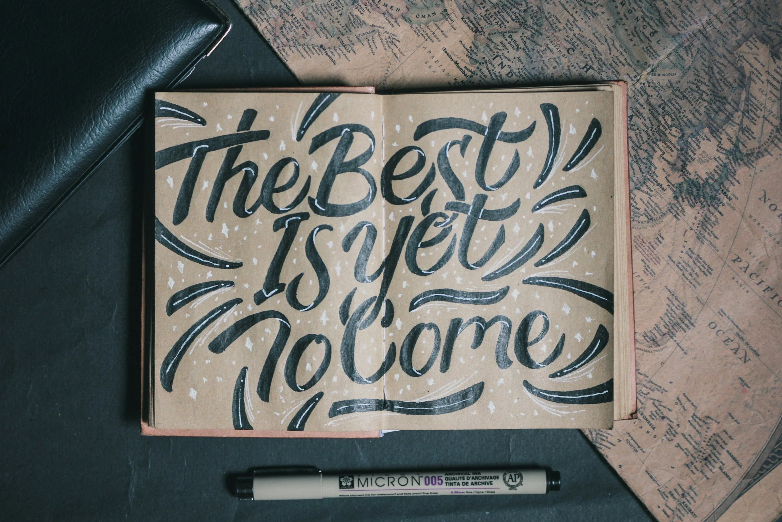 Close-up of a sketchbook with a handwritten message that says 'The best is yet to come' in black ink on a brown page, with a black marker underneath, surrounded by vintage maps and a black leather item.