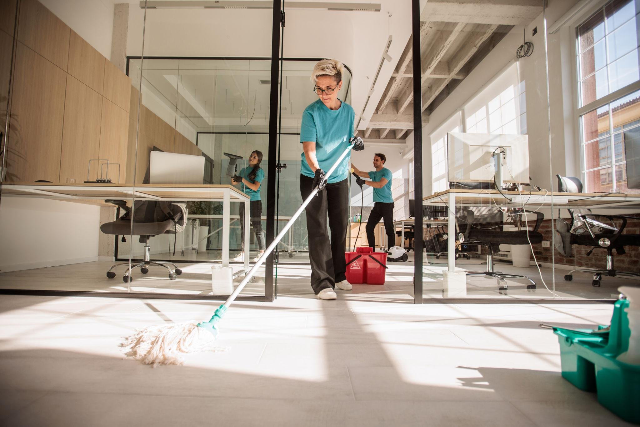 A woman cleaning the floor with a mop in an office space with large windows, desks, and computer monitors. Two other people are working in the background, and cleaning supplies are visible.