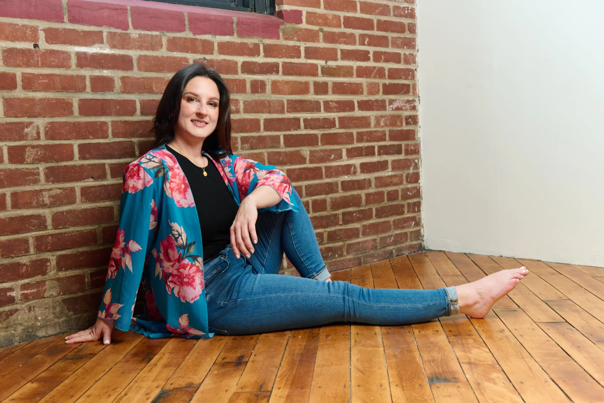 A woman with dark hair, wearing a black top and a colorful floral kimono, sitting on a wooden floor with her back against a brick wall, with her legs stretched out in front of her.