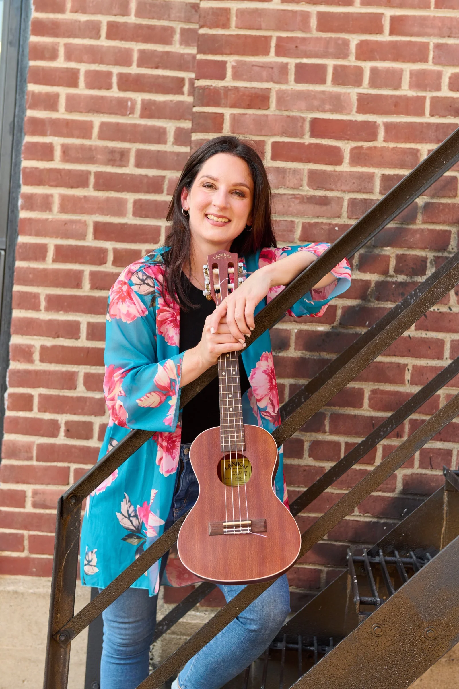 A young woman with dark hair, smiling, holding a small ukulele while leaning on a metal staircase railing against a brick wall background. She is wearing a vibrant blue floral cardigan.