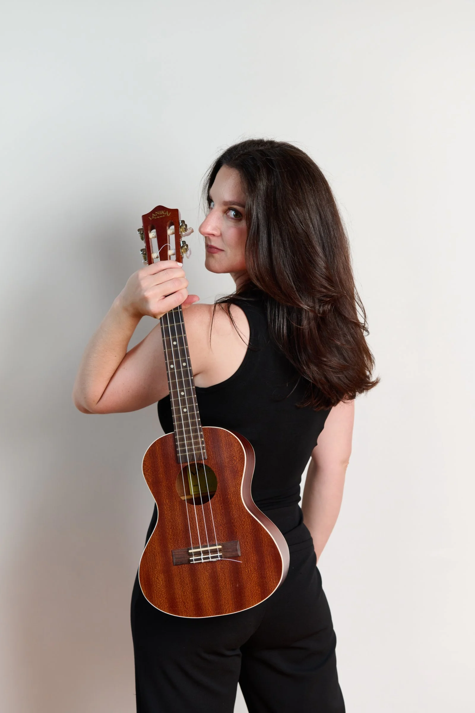 A woman with long brown hair in a black top holding a small wooden ukulele against a plain white wall.