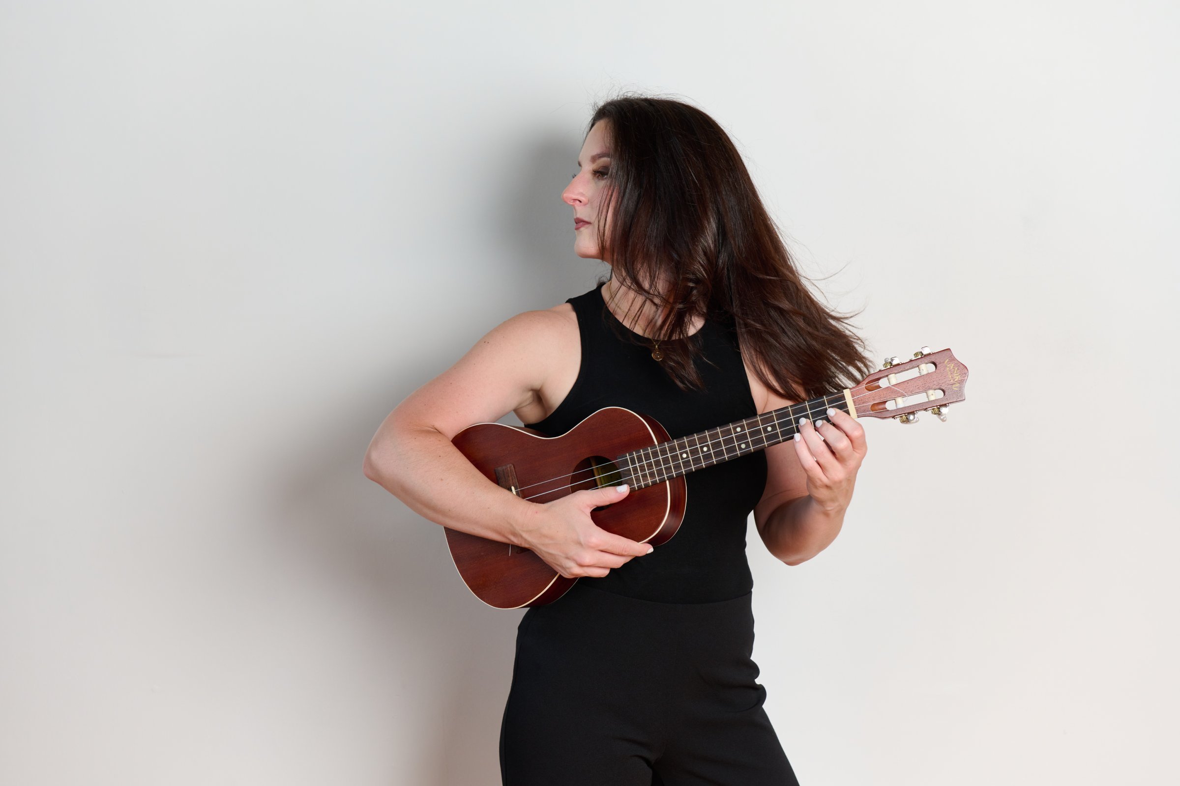 A woman with long brown hair wearing a black sleeveless top playing a ukulele against a plain white wall.