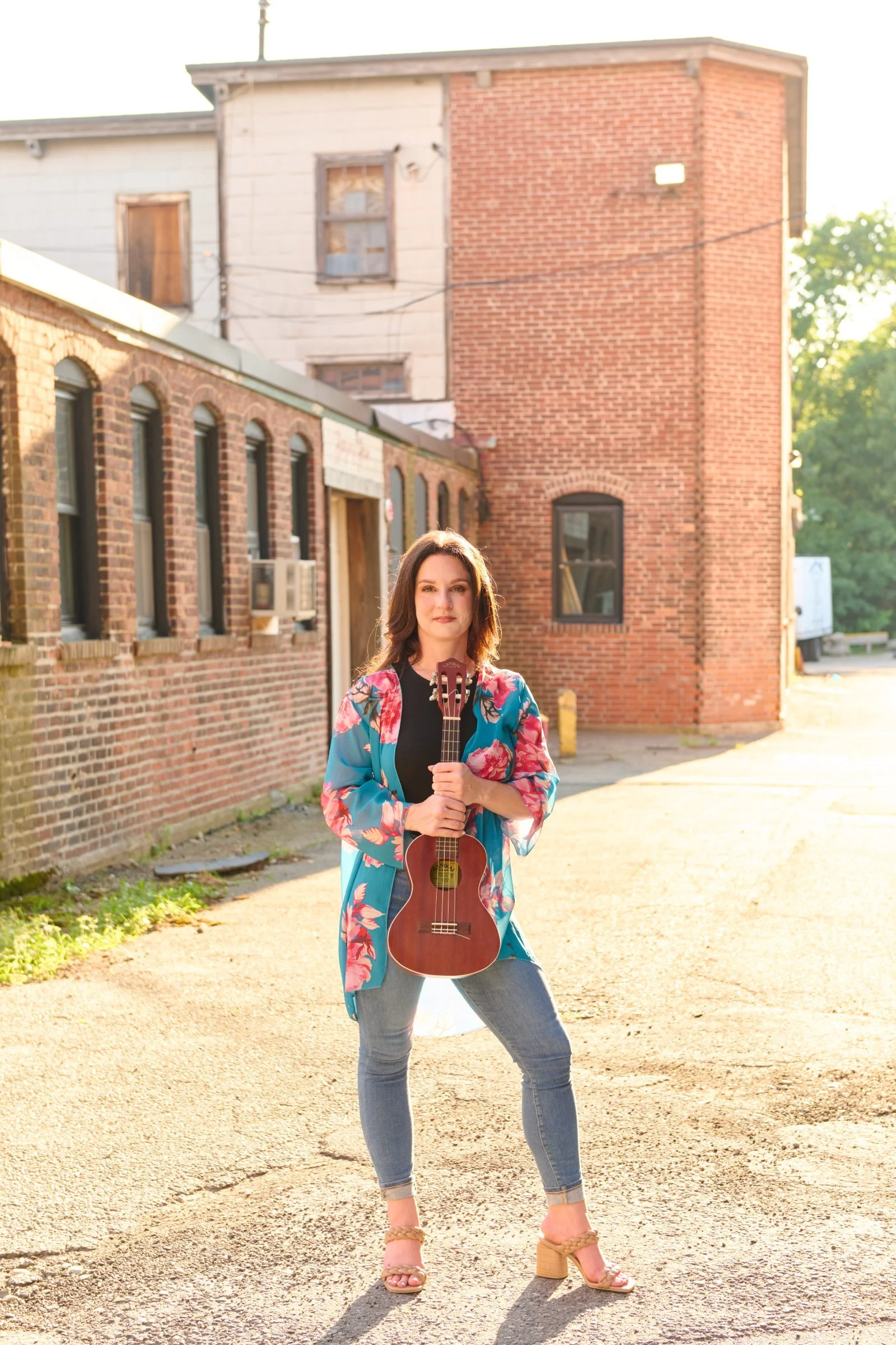 Woman standing outdoors on a sunny day holding a ukulele, wearing a floral jacket, jeans, and high-heeled sandals, in an urban alley with brick buildings in the background.