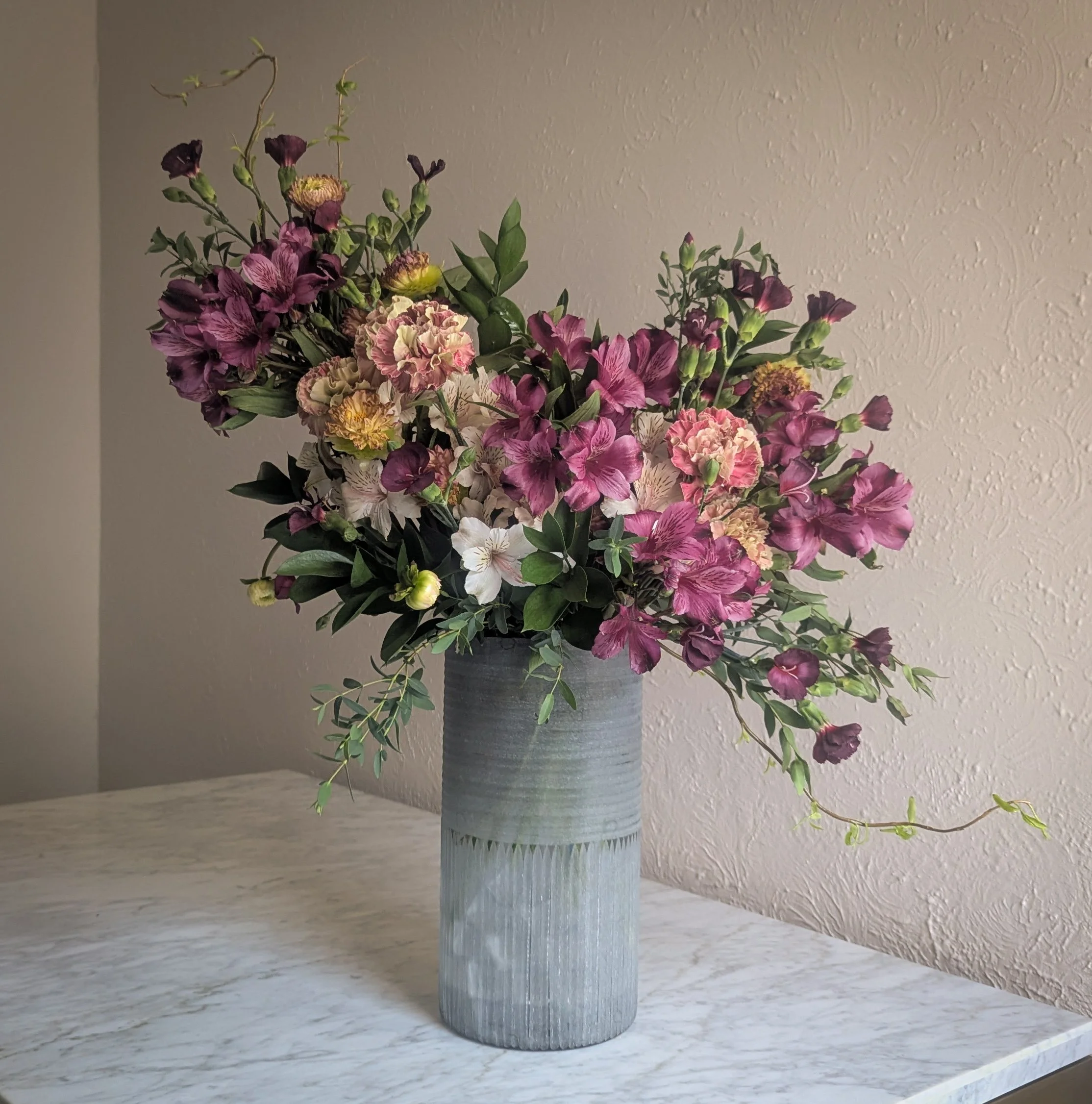 A thoughtfully designed floral arrangement featuring rich plum alstromeria, blush carnations, dahlias, and textured greenery in a contemporary glass vase. Created in Pinecone Florals' signature refined style for professional spaces.