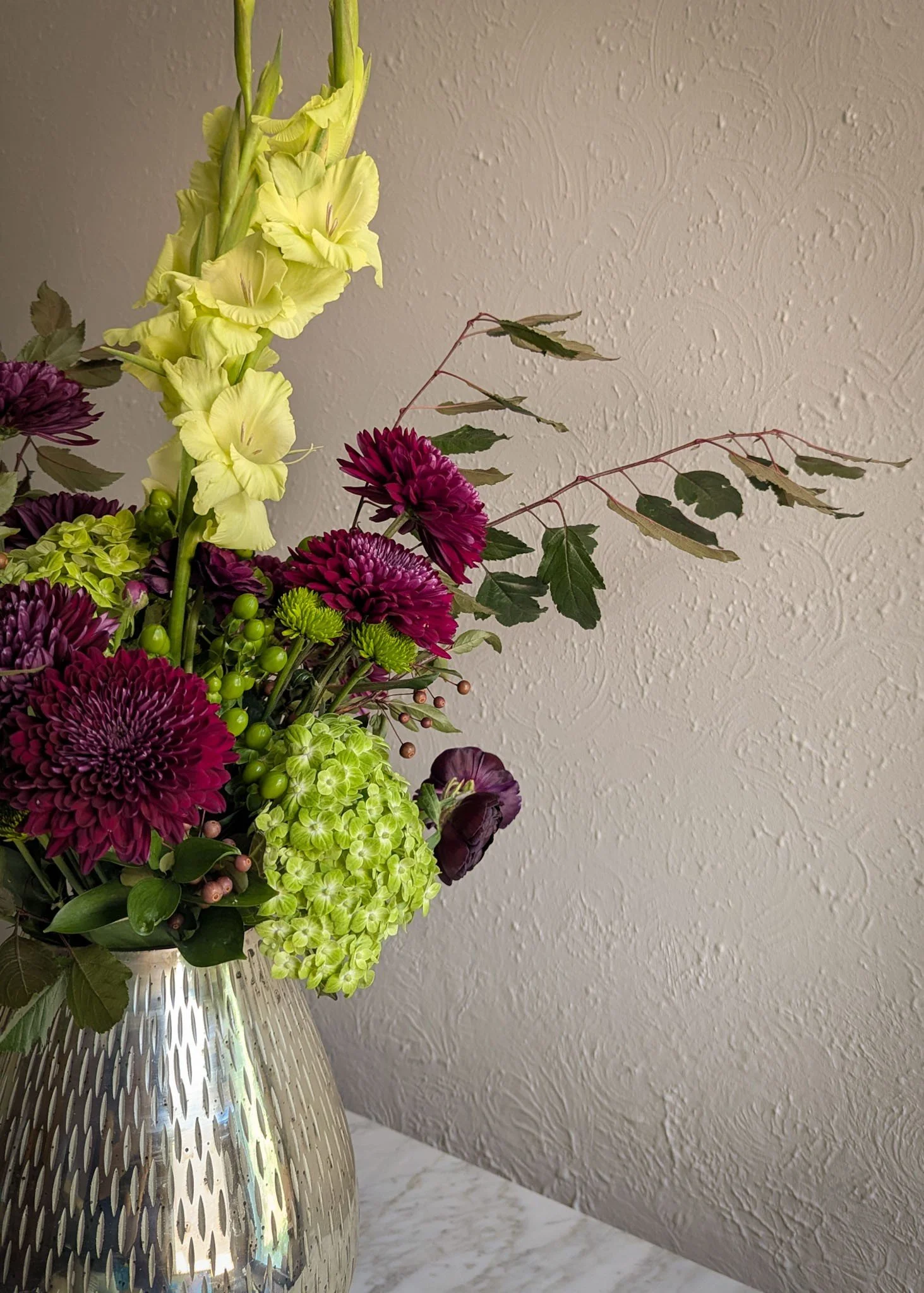 Sophisticated arrangement with rich cranberry mums, shamrock hydrangeas, gladiolas, and ranunculus in an elevated silver vase