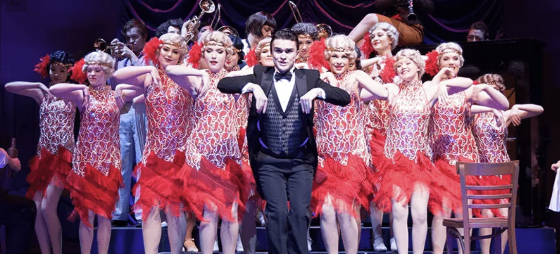 A theatrical stage performance with a man in a tuxedo and many women in red and white costumes, likely a musical or dance show.