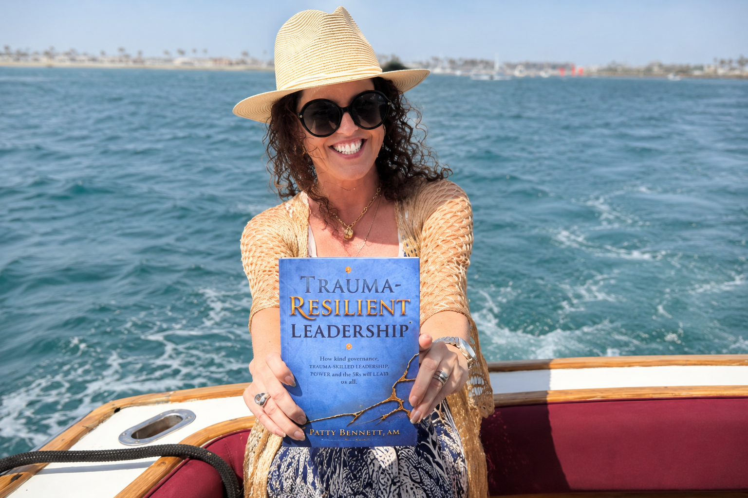 Patty with curly hair, wearing sunglasses, a straw hat, and a gold necklace, sitting on a boat, holding a book titled "Trauma-Resilient Leadership." She is smiling and enjoying a day on the water with a distant shoreline in the background.