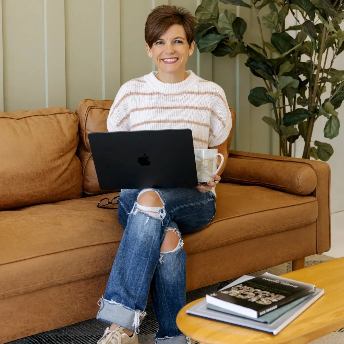 A woman sitting on a brown couch with ripped jeans and a white striped sweater, holding a laptop and a mug, smiling at the camera in a cozy living room with green plant and books on the table.