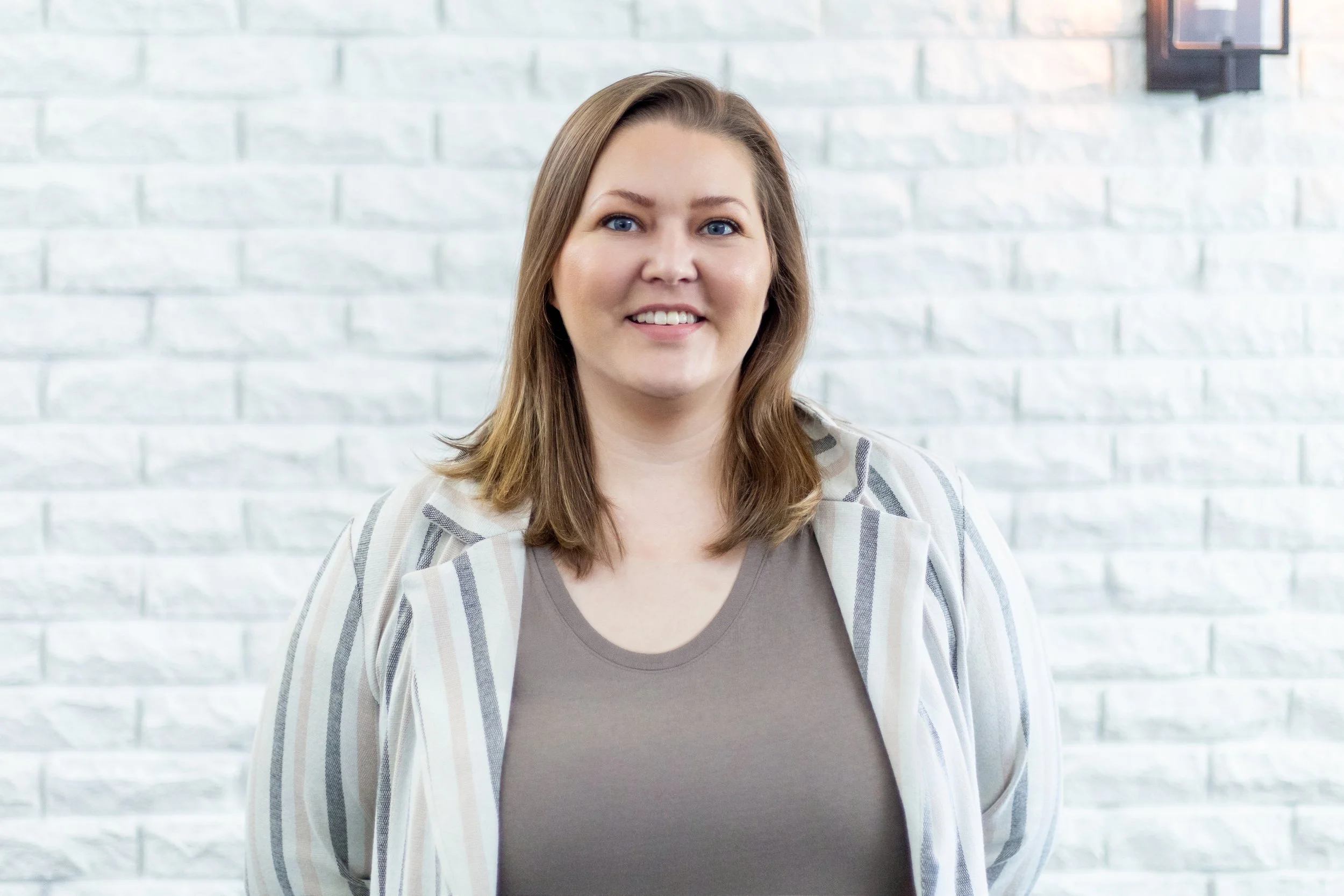 A woman with shoulder-length brown hair, blue eyes, and fair skin smiling in front of a white brick wall, wearing a striped blazer over a gray top.