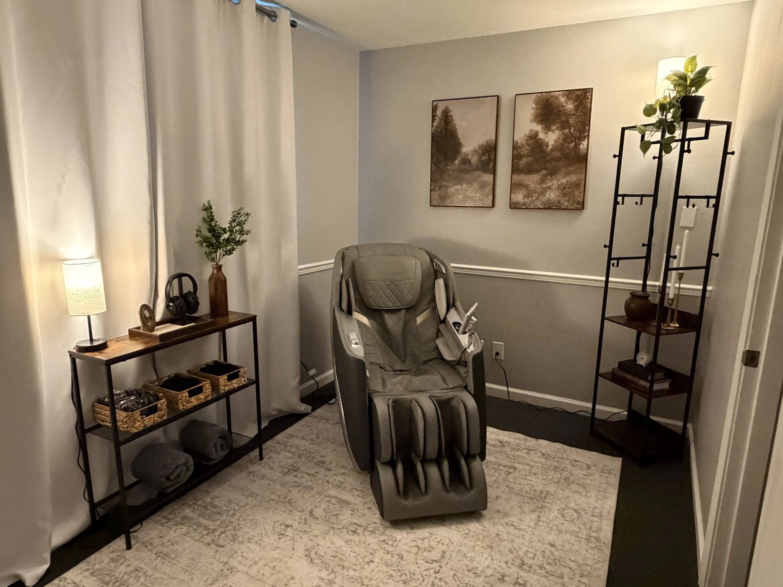A cozy interior room with gray walls, two framed landscape paintings, a standing black metal shelf with potted plant, small statue, candles, and a tall white curtain covering a window. There is a rocking chair with a padding, a small table with decor and baskets, a floor lamp, and a textured rug on the carpeted floor.