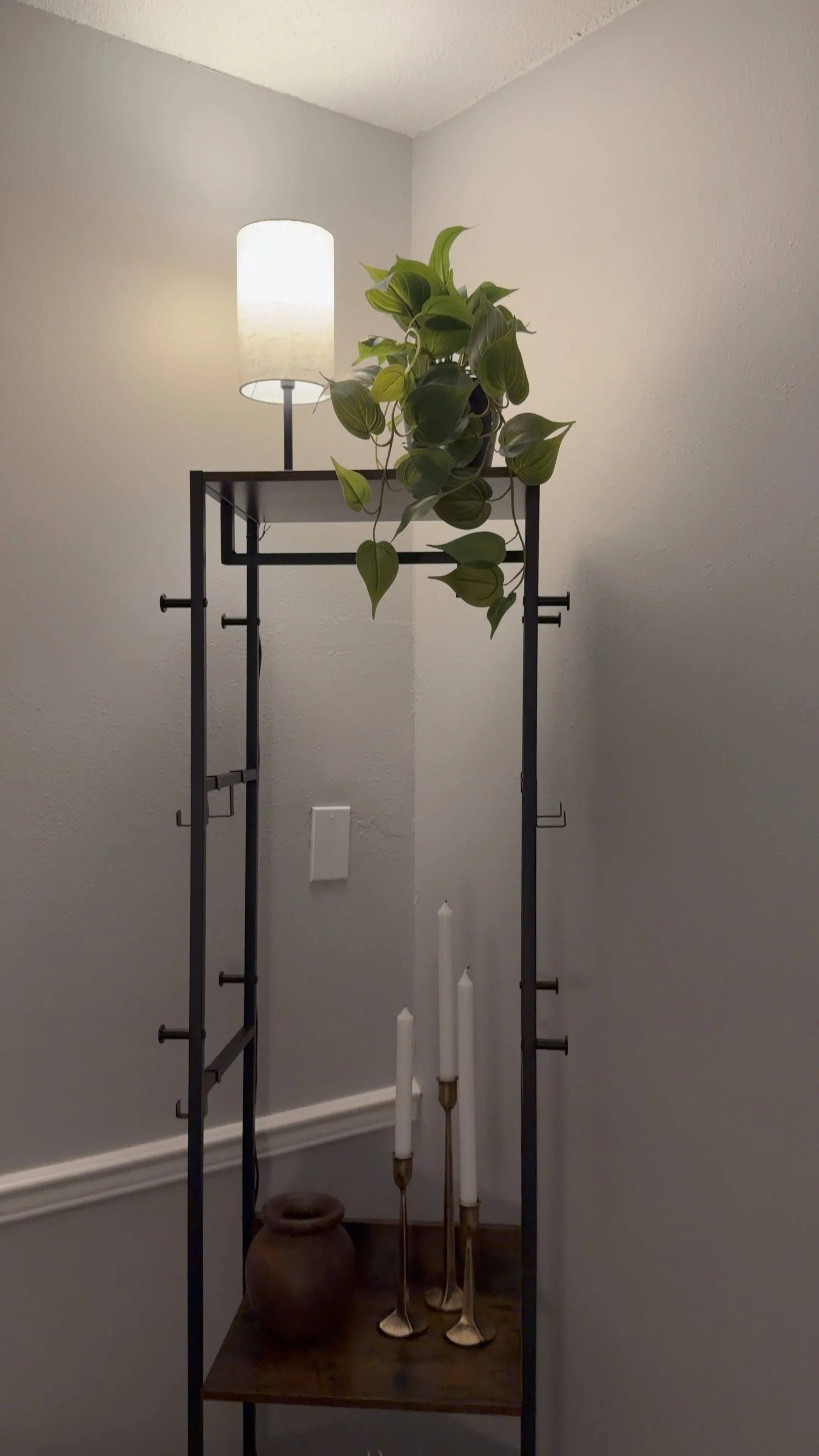 A black metal shelf with a white lampshade, a potted green plant, a brown vase, and three white candles in holders against a plain white wall.