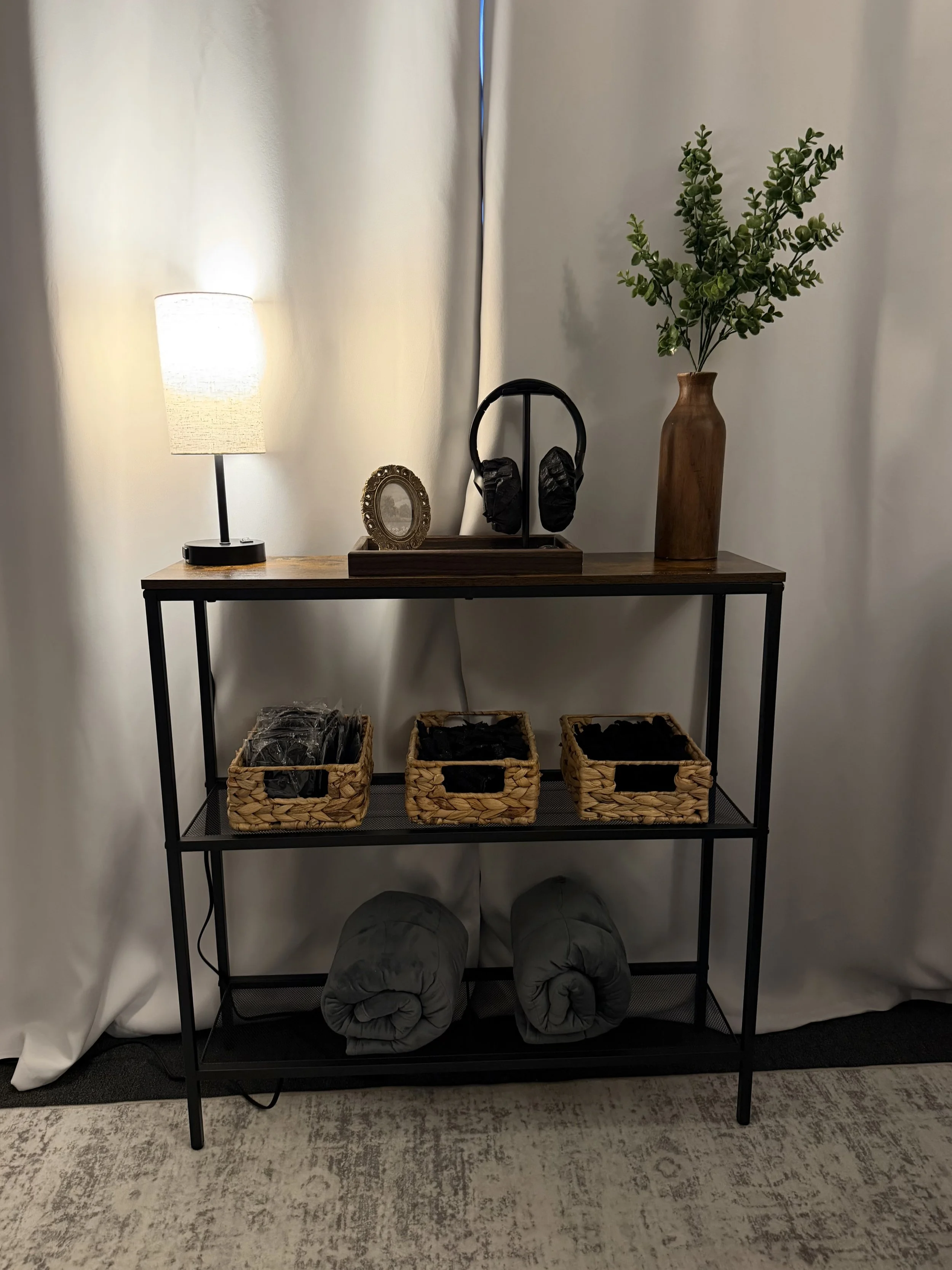 A black shelf with three baskets containing black items, three rolled-up gray camping mats underneath, a wooden vase with green foliage, a small ornate photo frame, a table lamp, and a headphone stand with black headphones on top, against white curtains.