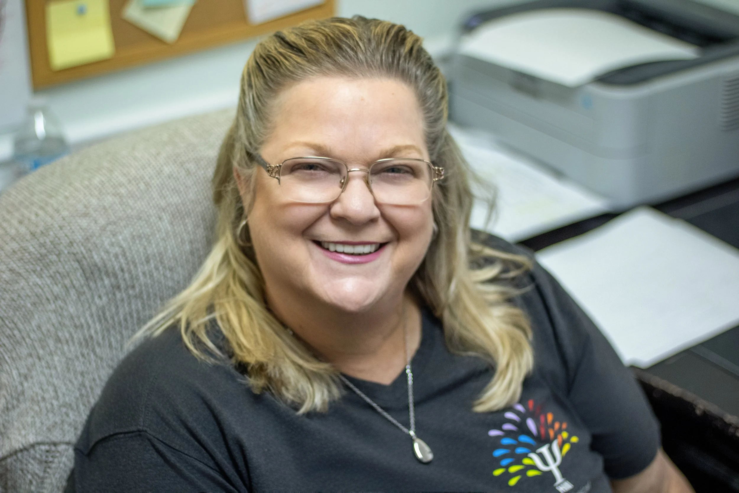 A woman with blonde hair, glasses, and a black T-shirt smiling while sitting in an office chair near a desk with papers and a printer.