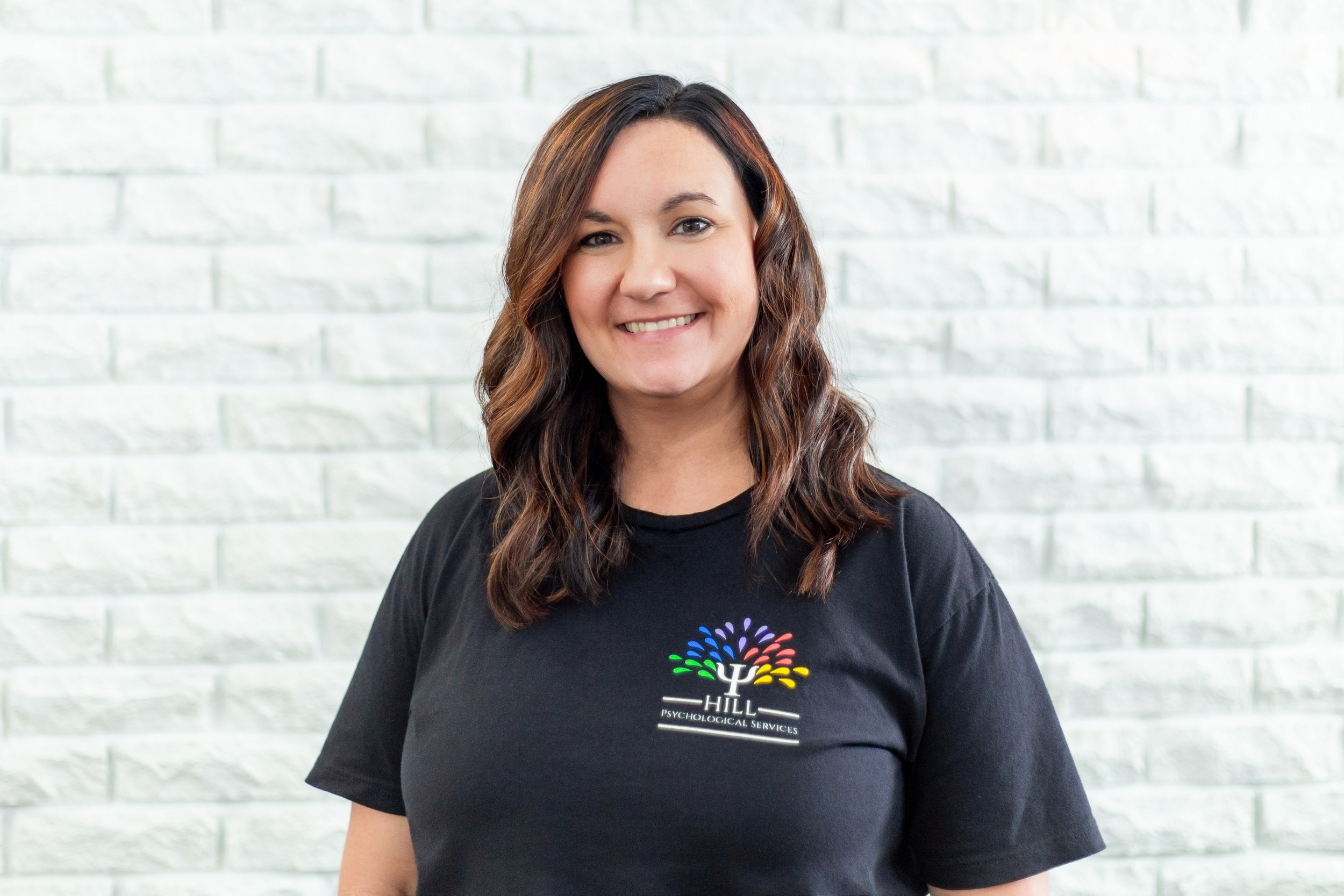A woman with long wavy brown hair smiling, wearing a black T-shirt with a colorful logo on it, standing in front of a light-colored brick wall.