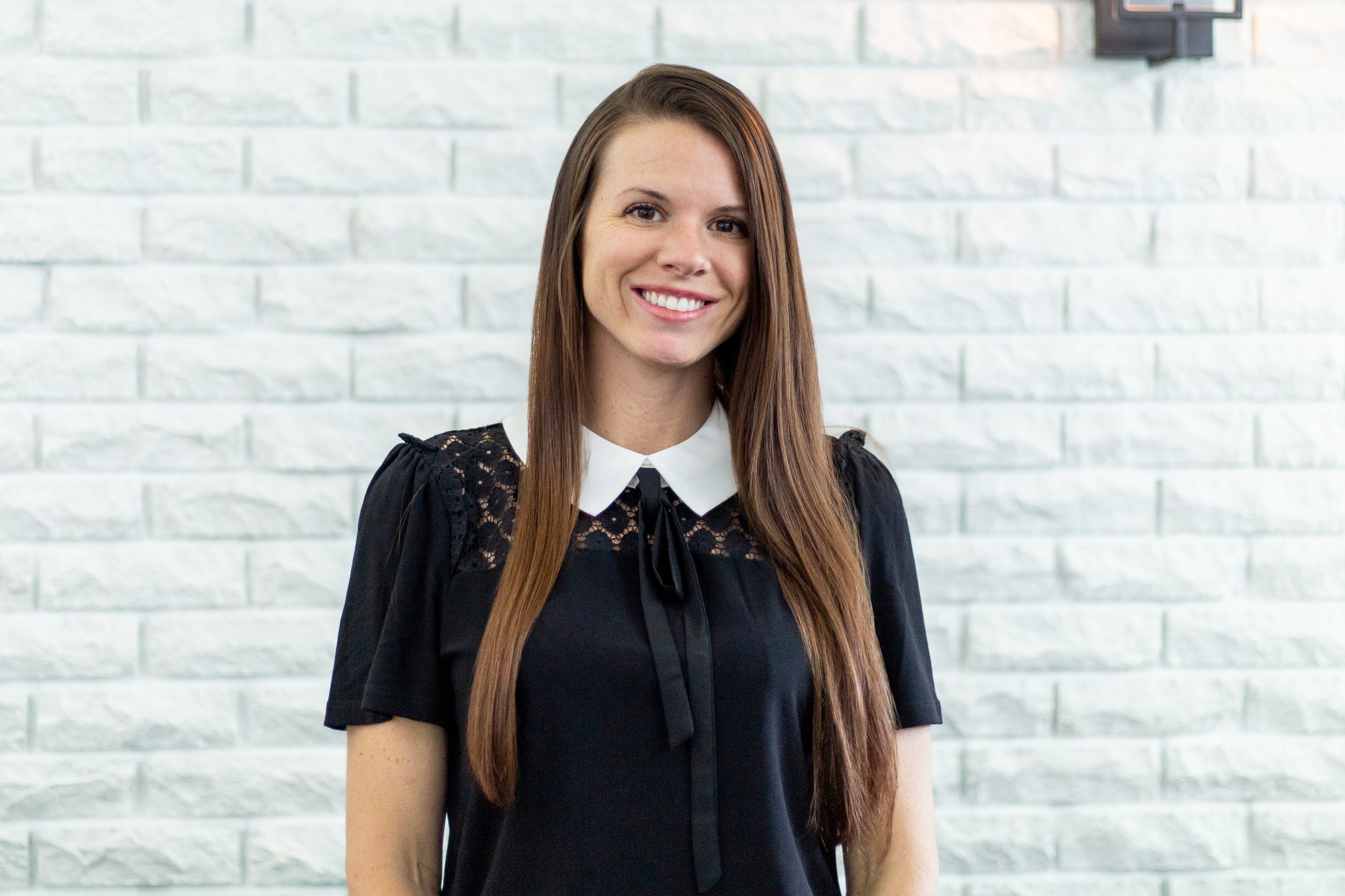 A young woman with long brown hair smiling, wearing a black dress with lace details and a white collar, standing in front of a white brick wall.