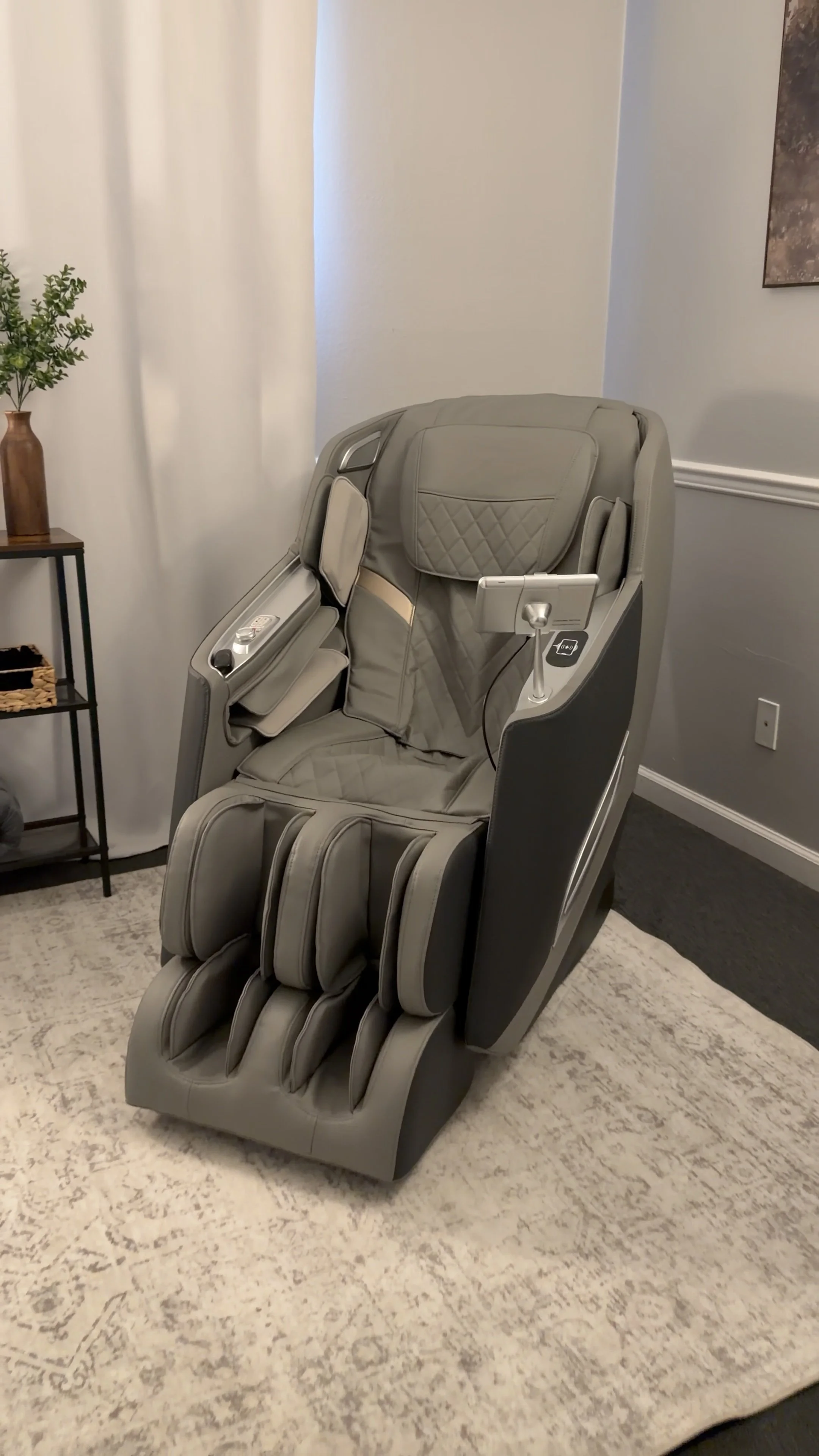 A beige massage chair with multiple padded sections, armrests, and control panel in a room with a patterned area rug, a black side table with a plant, and framed wall art.