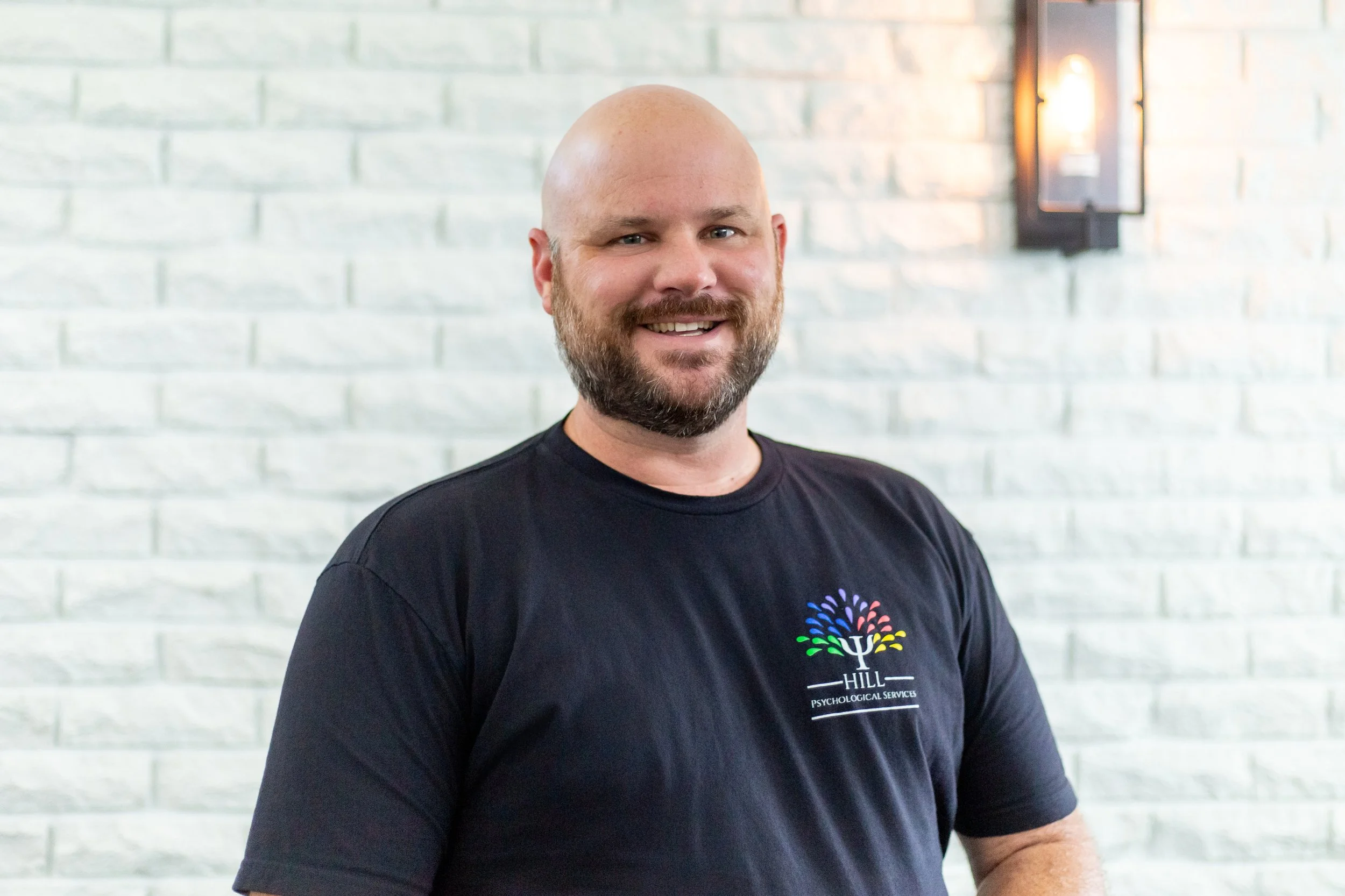 A man with a shaved head and beard smiling, wearing a black T-shirt with a colorful tree logo and text, standing in front of a light-colored brick wall with a wall-mounted light fixture.