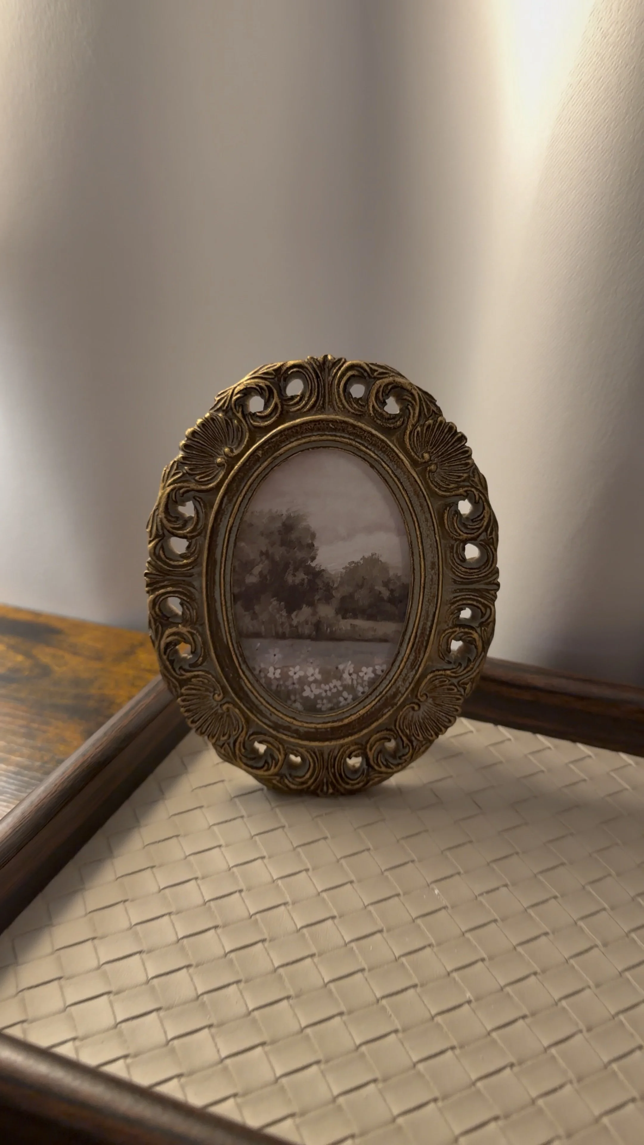 A decorative oval picture frame with a landscape painting inside, resting on a beige woven surface, with a woven basket and a tray with handles in the background.