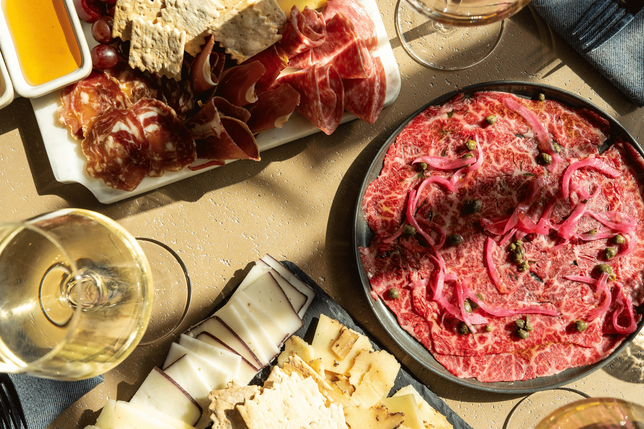 A charcuterie board with assorted cheeses, cured meats, honey, and sliced crackers, alongside a glass of white wine and a dish of raw beef topped with pink onions and capers.