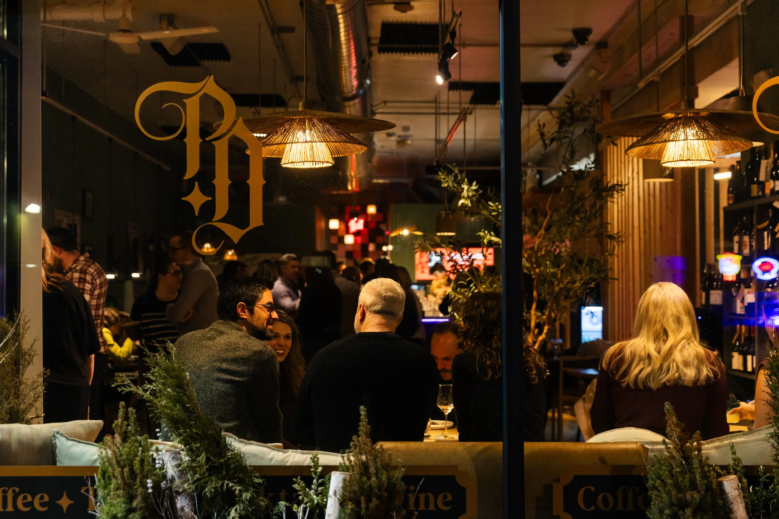 A lively indoor restaurant scene seen through a window with yellow lettering. People are gathered around tables, engaging in conversation, with warm lighting and hanging lamps creating a cozy atmosphere. There is greenery visible in the foreground.