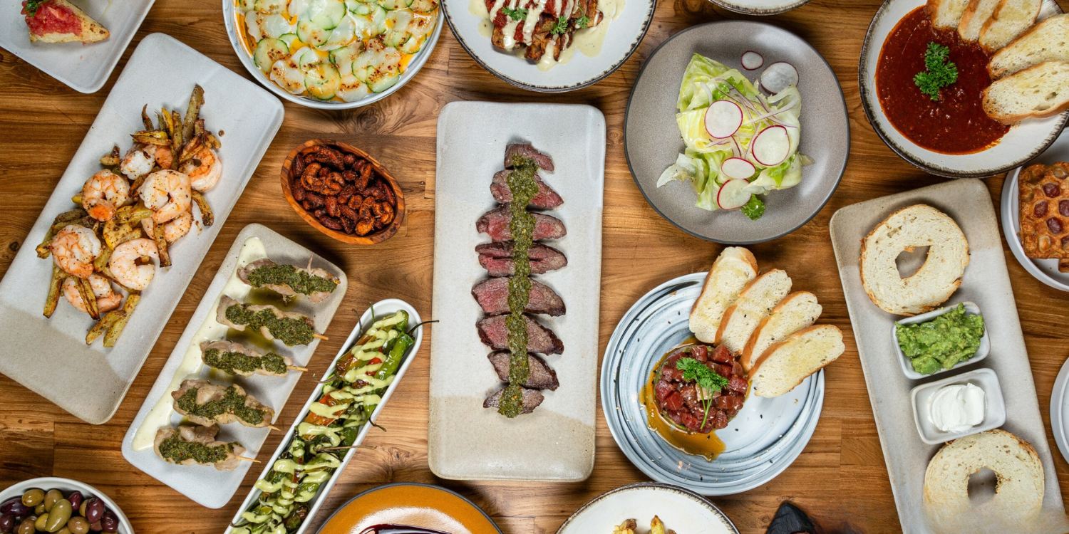 Assorted dishes on a wooden table including grilled shrimp, sliced steak with green sauce, a salad with radish slices, bread with dip, shrimp skewers, and various appetizers and sides.