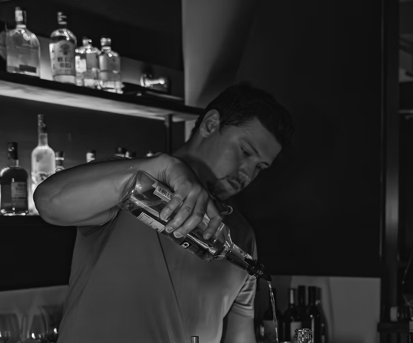 A man pouring a drink from a bottle into a glass behind a bar with liquor bottles on shelves in the background.