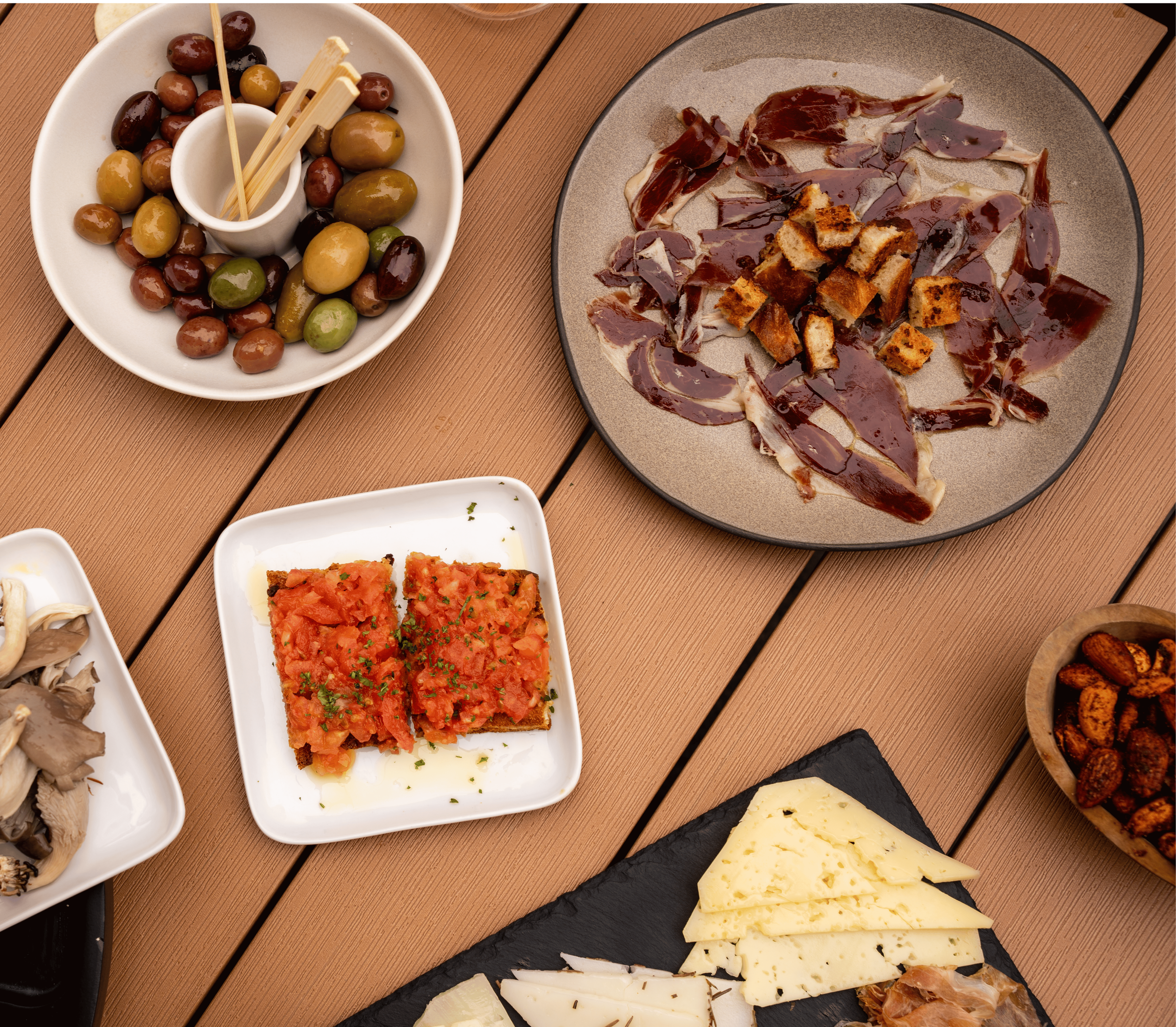 Assorted dishes of food on a wooden table, including a plate of mixed olives, a platter of sliced cured meats with sauce, a white dish with tomato-based dish, and a black slate with various cheeses.