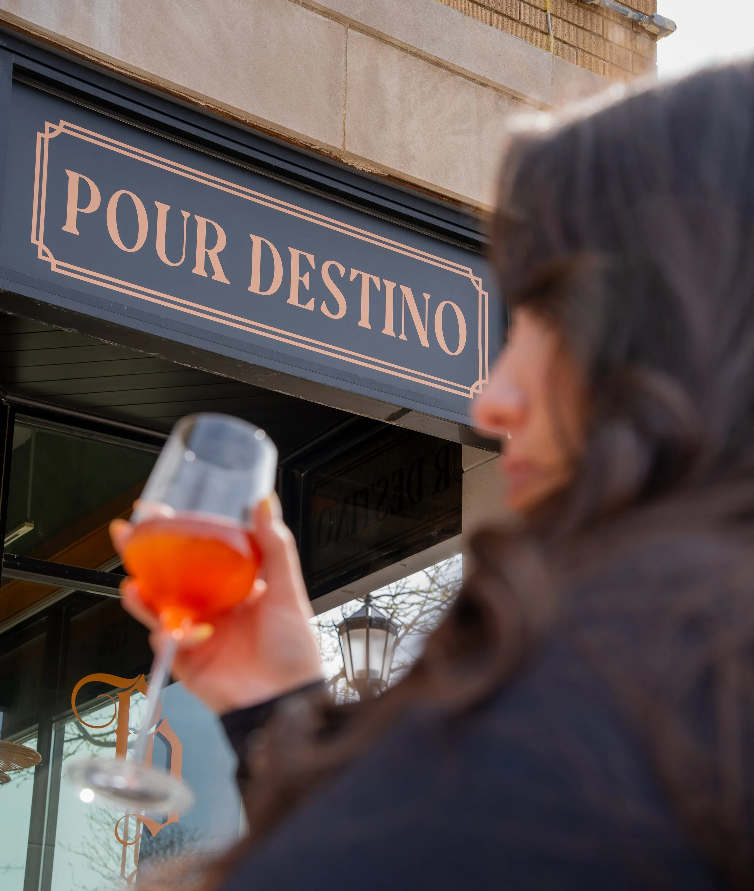 A person holding a glass of orange-colored beverage in front of a store sign that reads "POUR DESTIN" with the person's face partially visible and blurred; the image is taken outdoors.
