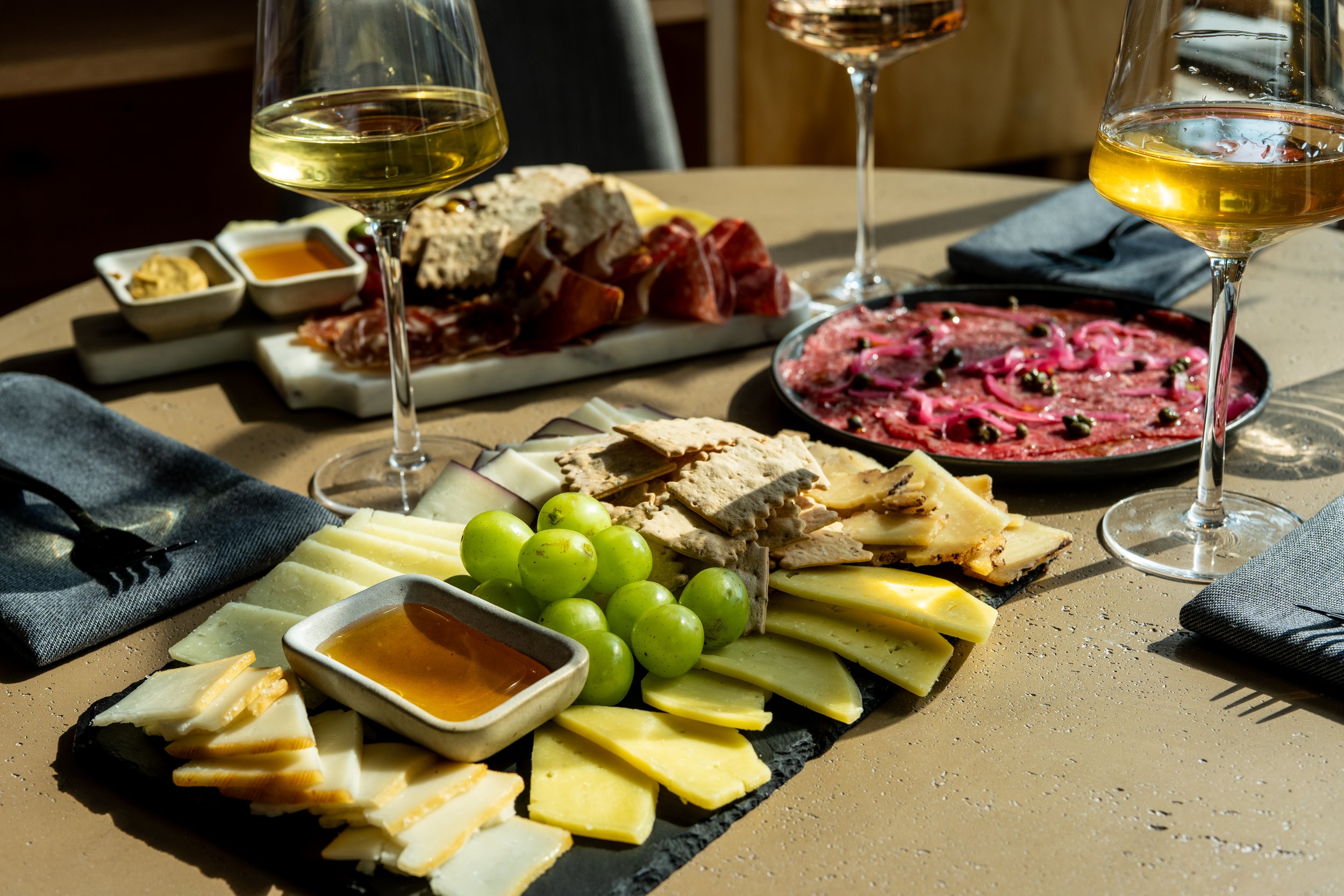 A charcuterie board with a variety of cheeses, green grapes, sliced meats, crackers, honey, and dipping sauces, with two glasses of white wine on a table.