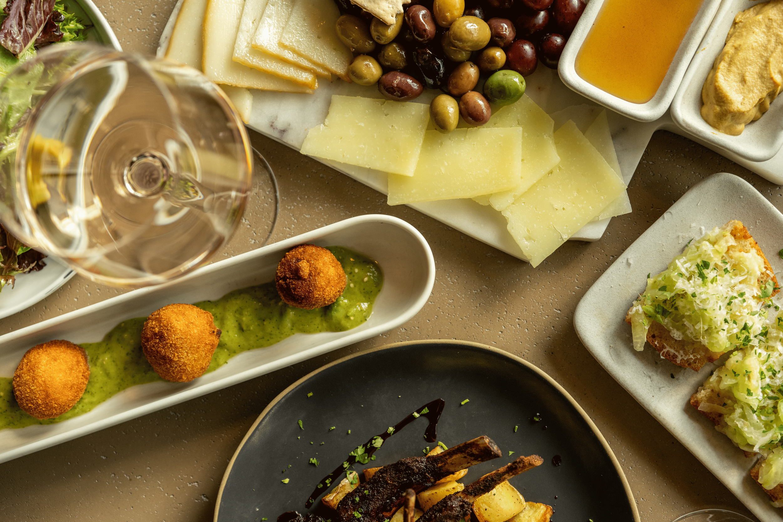 A variety of appetizers on white and black plates, including cheese, olives, salad, croquettes with green sauce, and grilled vegetables, along with a glass of white wine on a beige textured table.