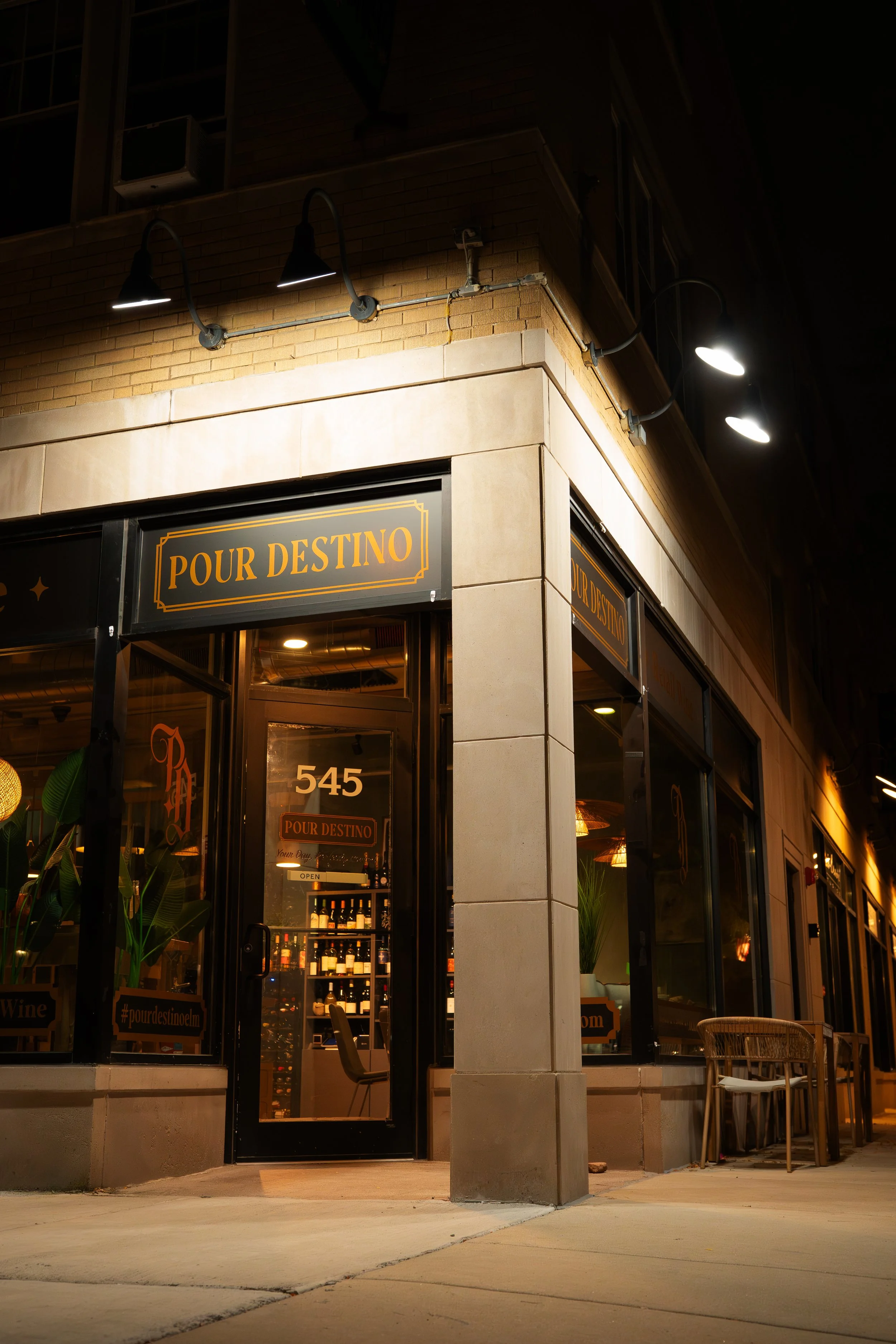 The exterior of a restaurant named Pour Destino, with a black sign with gold lettering, large glass windows showing the interior, and outdoor seating including chairs and tables, at night time illuminated by overhead lights.