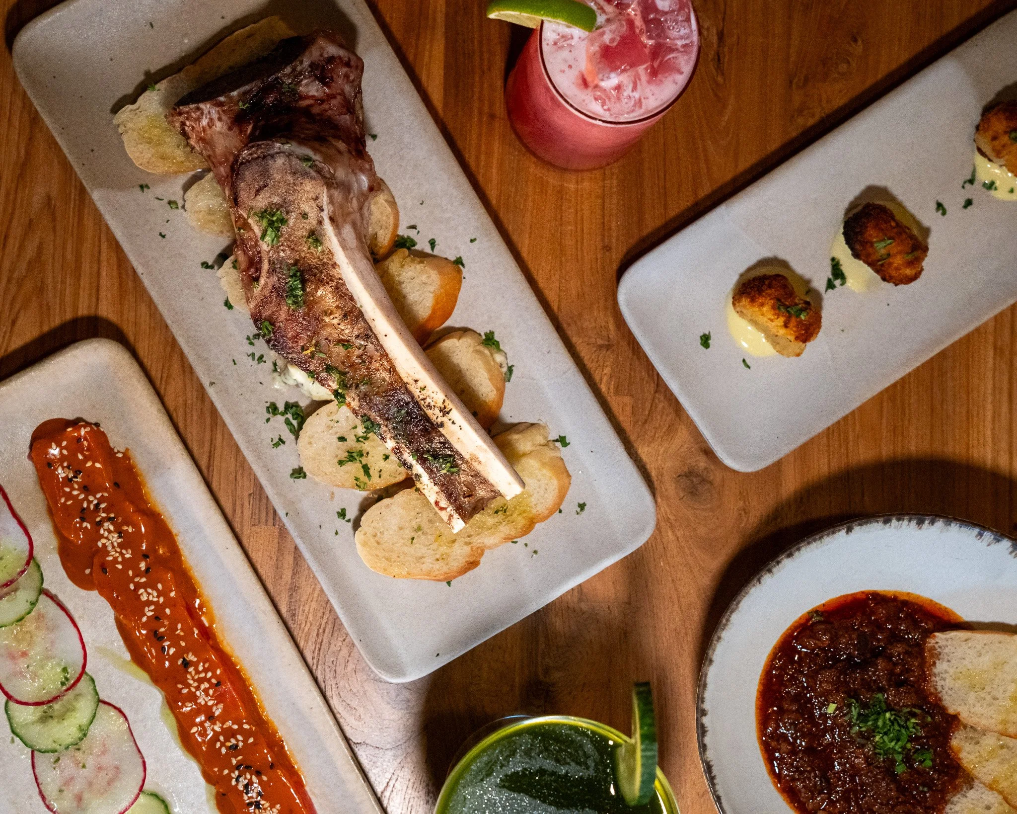 assortment of dishes on a wooden table, including a grilled piece of meat with lemon slices and herbs, three pieces of fried food with sauce, and various dipping sauces and garnishes.