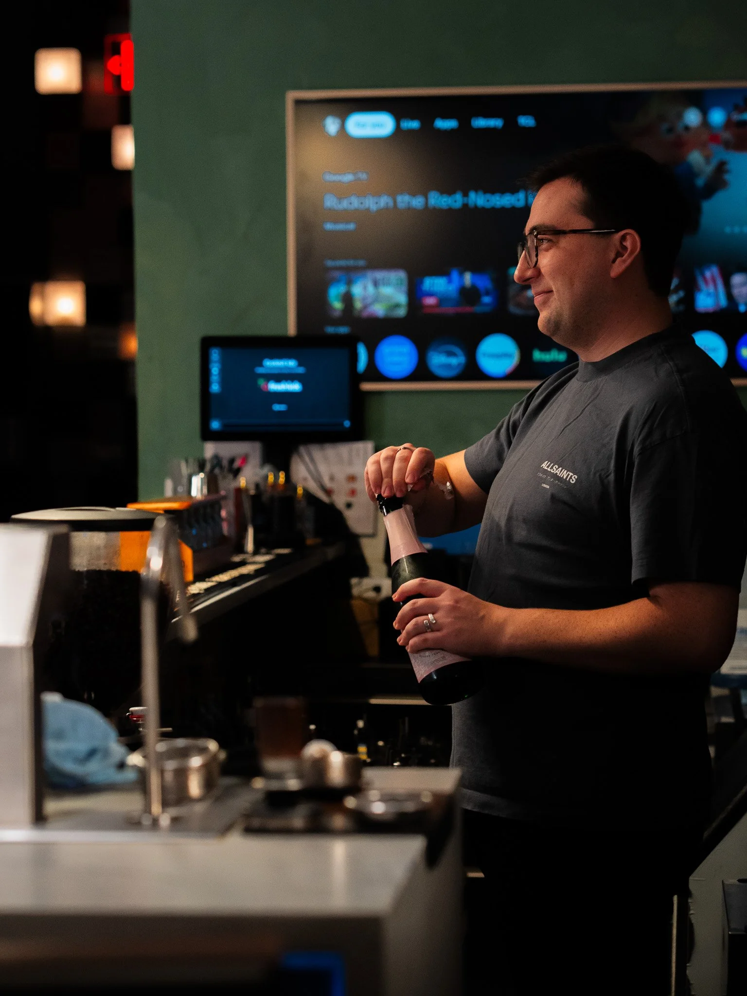 A man with glasses wearing a 'ALLSAINTS' t-shirt smiles while holding a bottle of champagne in a restaurant or bar setting, with a large TV screen and a smaller monitor in the background, along with some kitchen or bar equipment in the foreground.