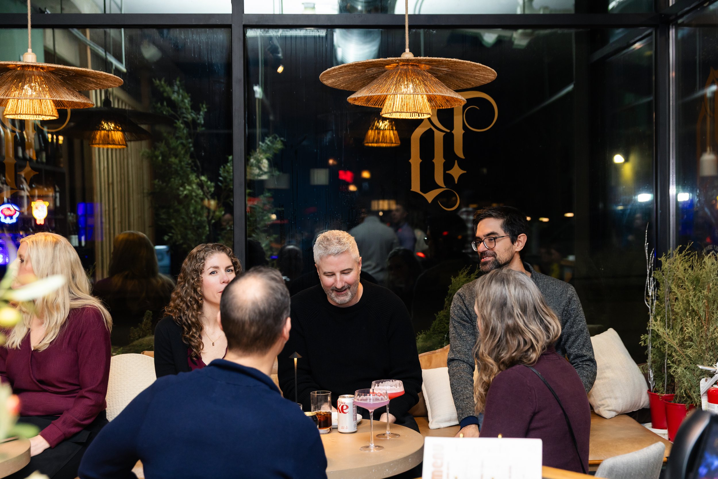 Group of people gathered around a table at a restaurant, socializing and enjoying drinks, with decorative wooden pendant lights hanging from the ceiling and large windows in the background.
