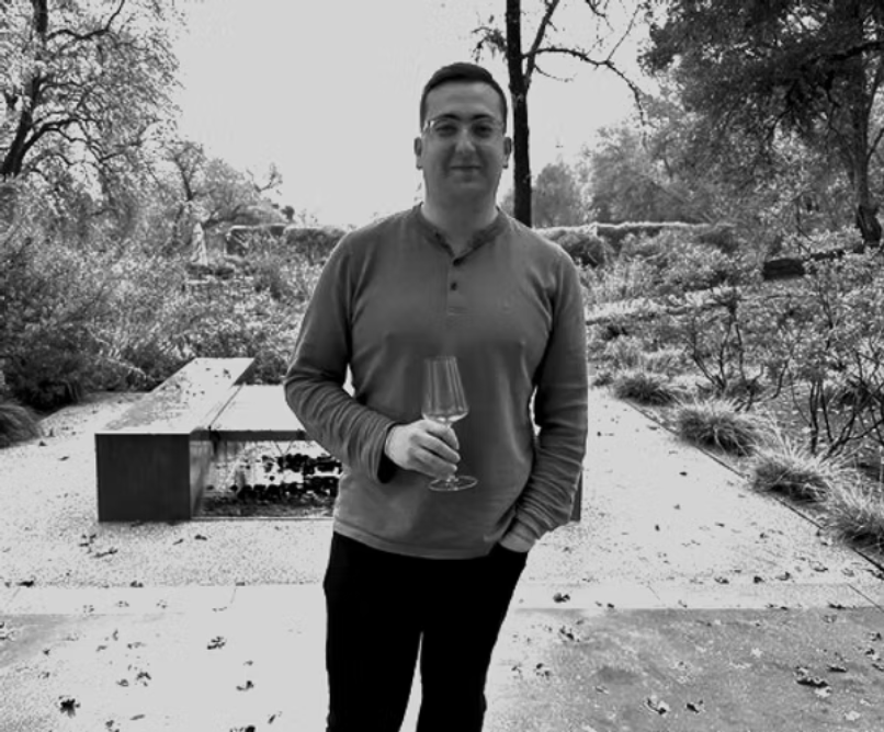 A man holding a wine glass standing outdoors in a garden or park with trees and plants, in black and white.