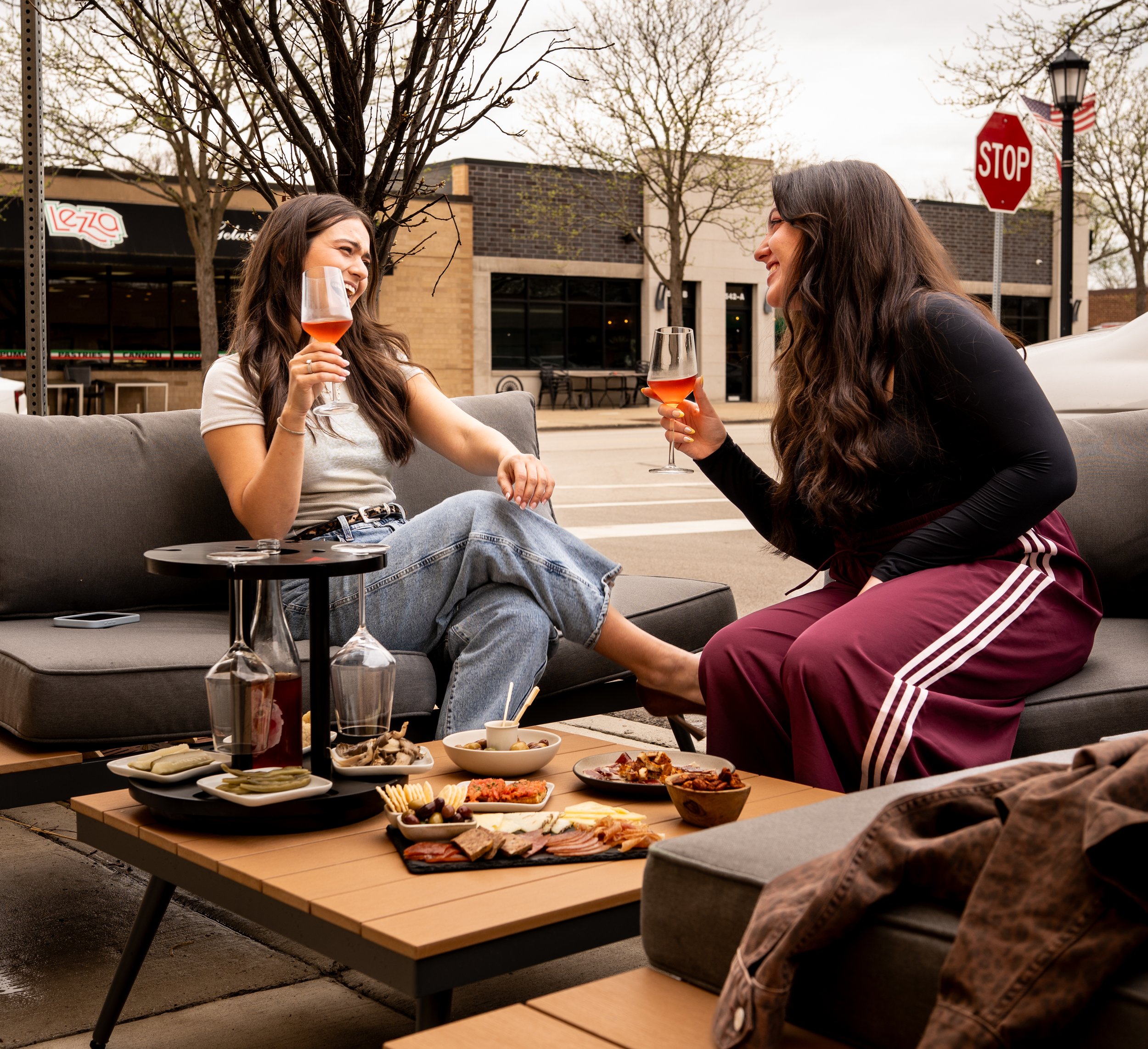 Two women enjoying drinks and conversation outdoors on a city sidewalk with a table of assorted snacks.