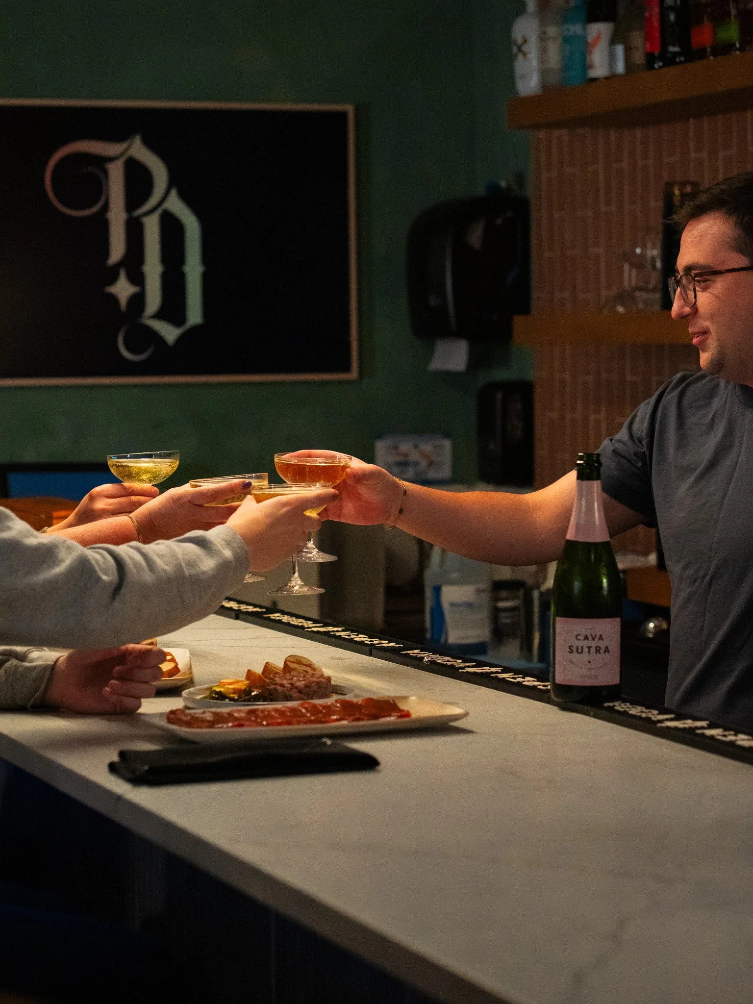 People enjoying drinks and appetizers at a bar or restaurant with a person serving wine and a bottle of cava on the counter.