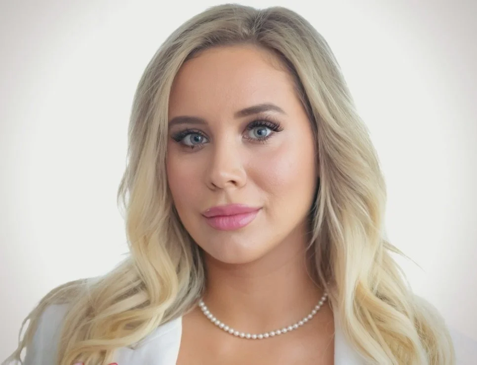 A woman with long, wavy blonde hair, blue eyes, and pink lipstick, wearing a white top and a pearl necklace, facing the camera with a slight smile.